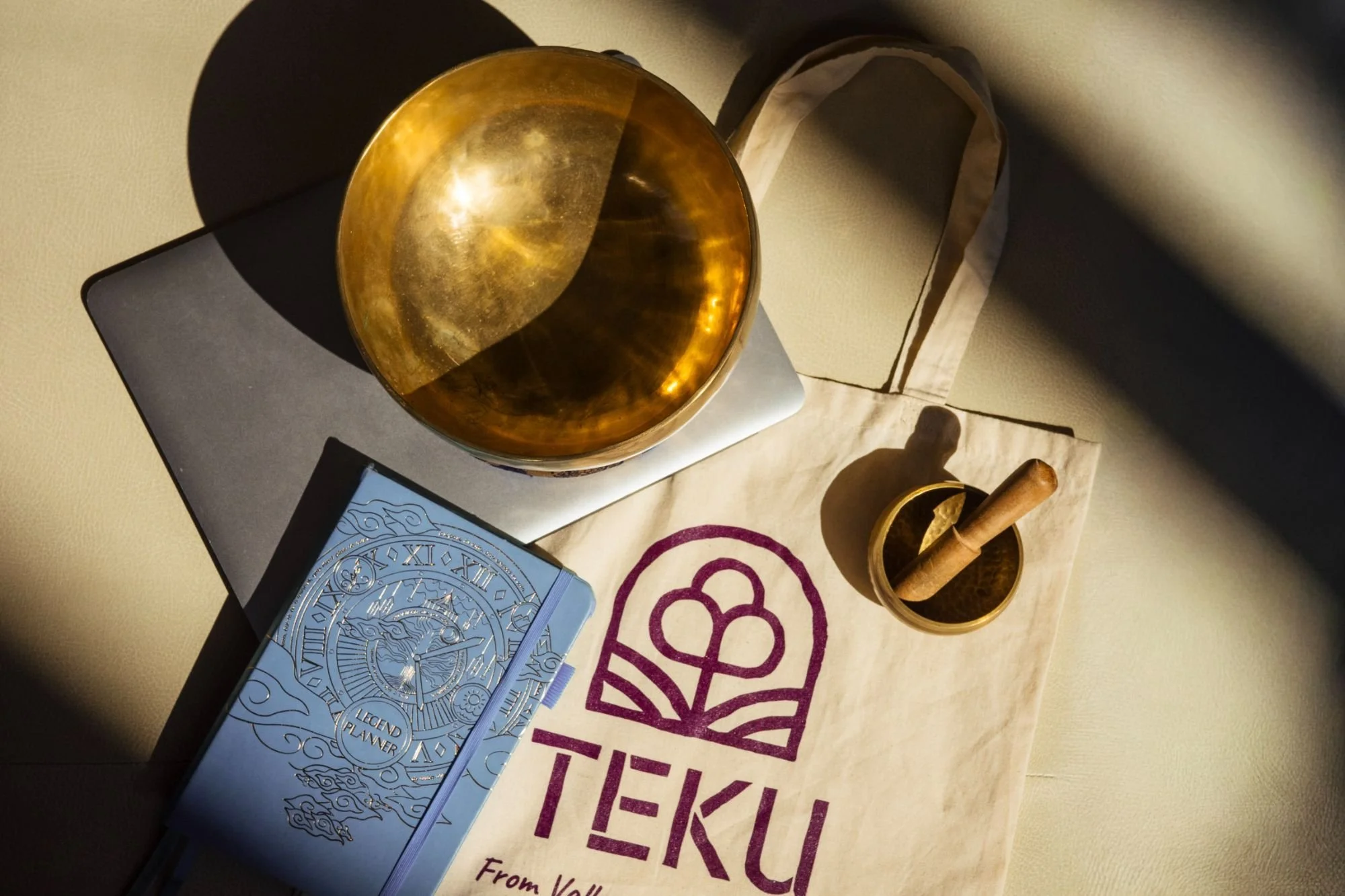 A flat lay of a gold bowl, a blue book titled 'Legend Planner', a paper bag with the word 'TEKU' and a logo, a small mortar and pestle, and a metallic scale on a beige surface with a shadow cast by sunlight.