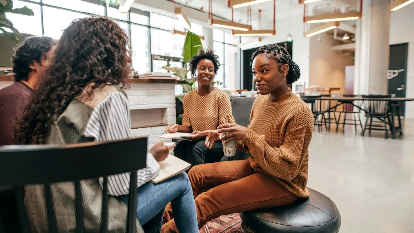 Group of four women sitting in a modern cafe, engaged in conversation. The woman on the right holds a disposable coffee cup. The cafe has large windows, plants, and contemporary decor.