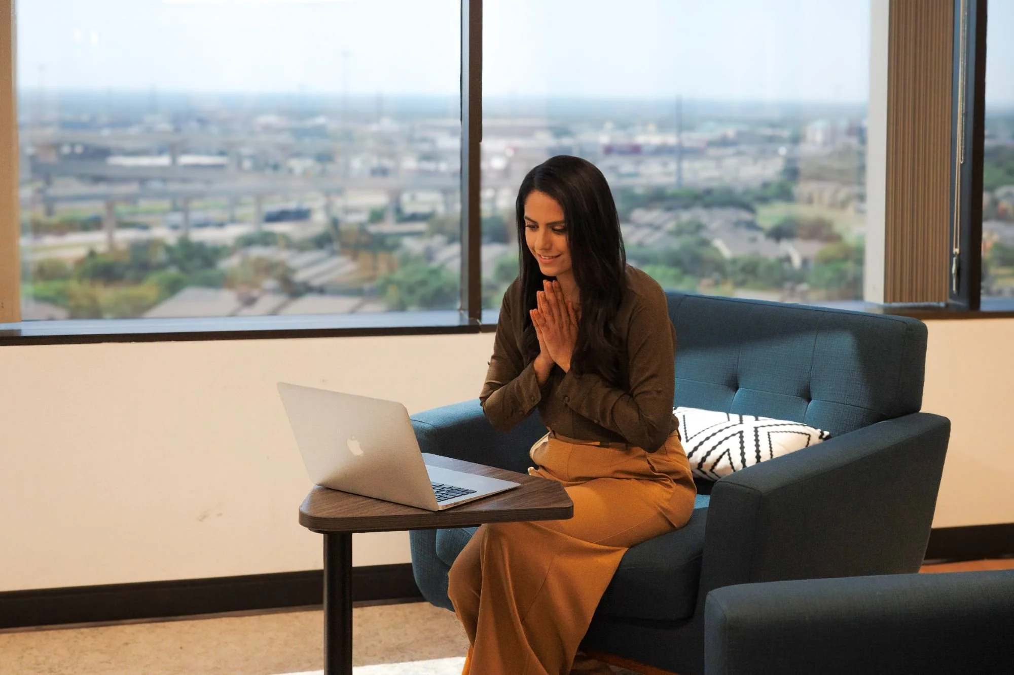 A woman sitting on a blue sofa with a laptop on a small wooden table in front of her, hands clasped together, smiling, with large windows showing cityscape views.