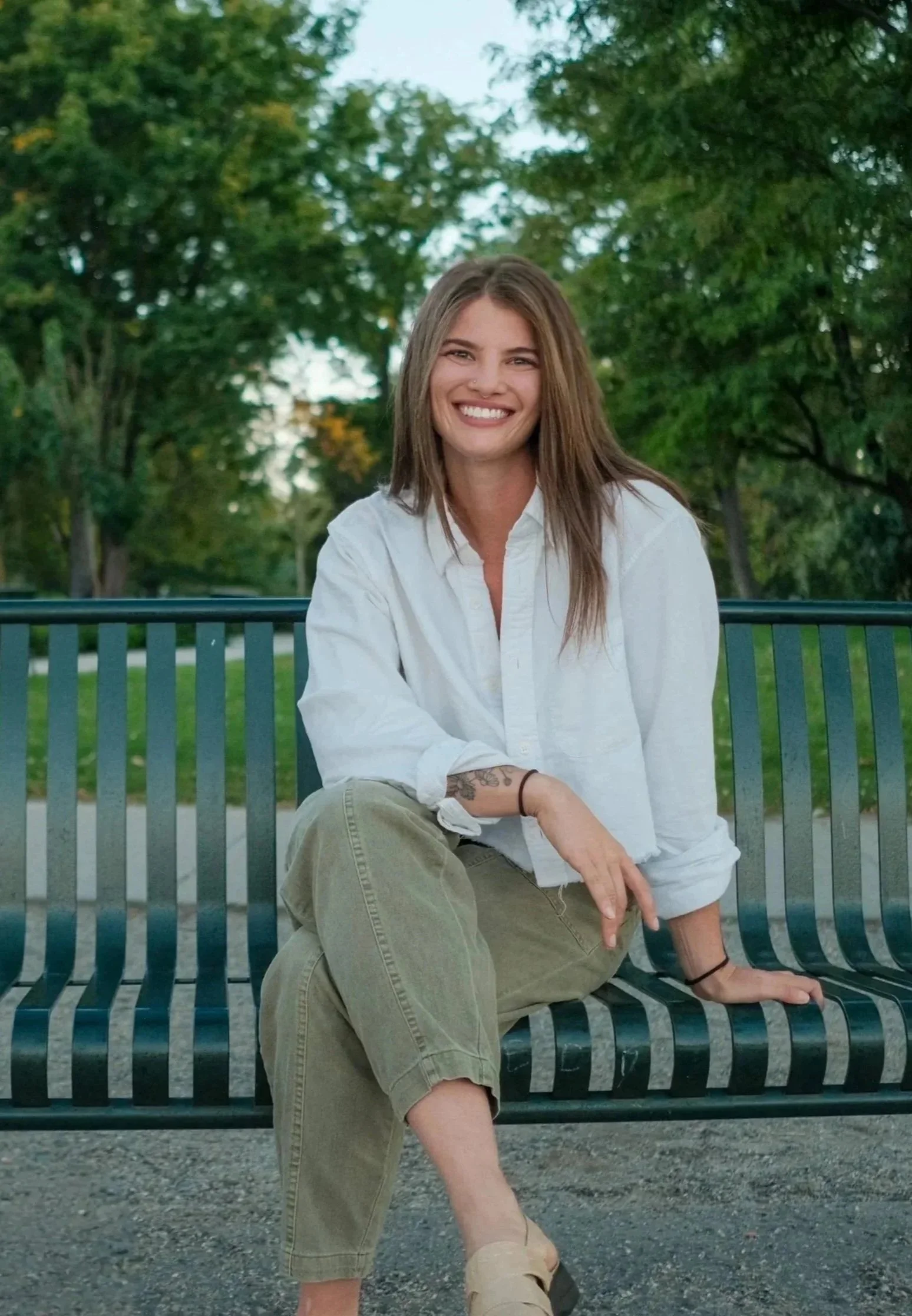 A smiling woman with long brown hair sitting on a black metal park bench, wearing a white button-up shirt, khaki pants, and beige slip-on shoes, outdoors with green trees in the background.