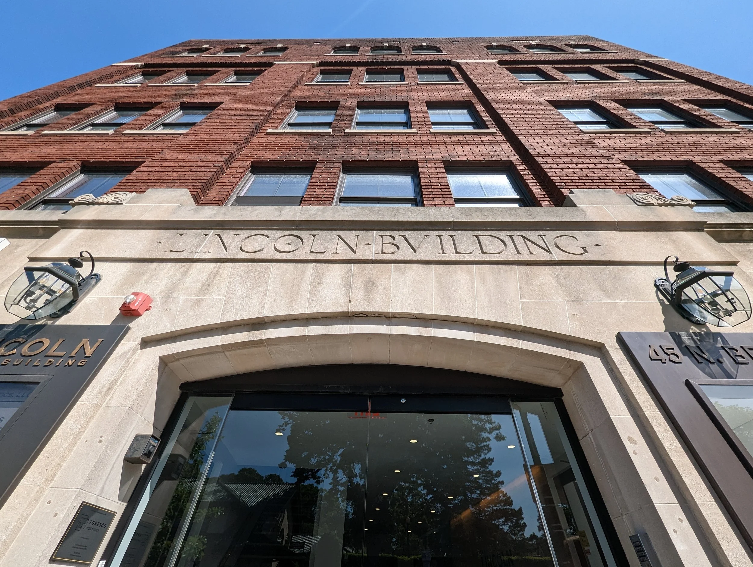 The Lincoln Building, a red brick multi-story structure with a stone entrance and large glass doors, is shown from a low angle against a clear blue sky, with lantern-style lights on either side of the entrance.