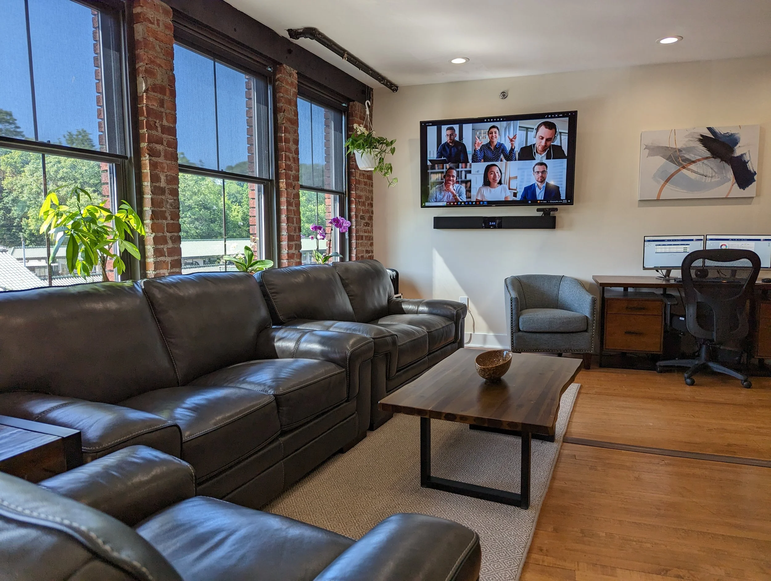 Office lounge with large leather sofa, potted plants on windowsill, television displaying a video conference, wooden coffee table with decorative bowl, gray armchair, desk with dual monitors, and artwork on the wall, with exposed brick and large windows letting in natural light.