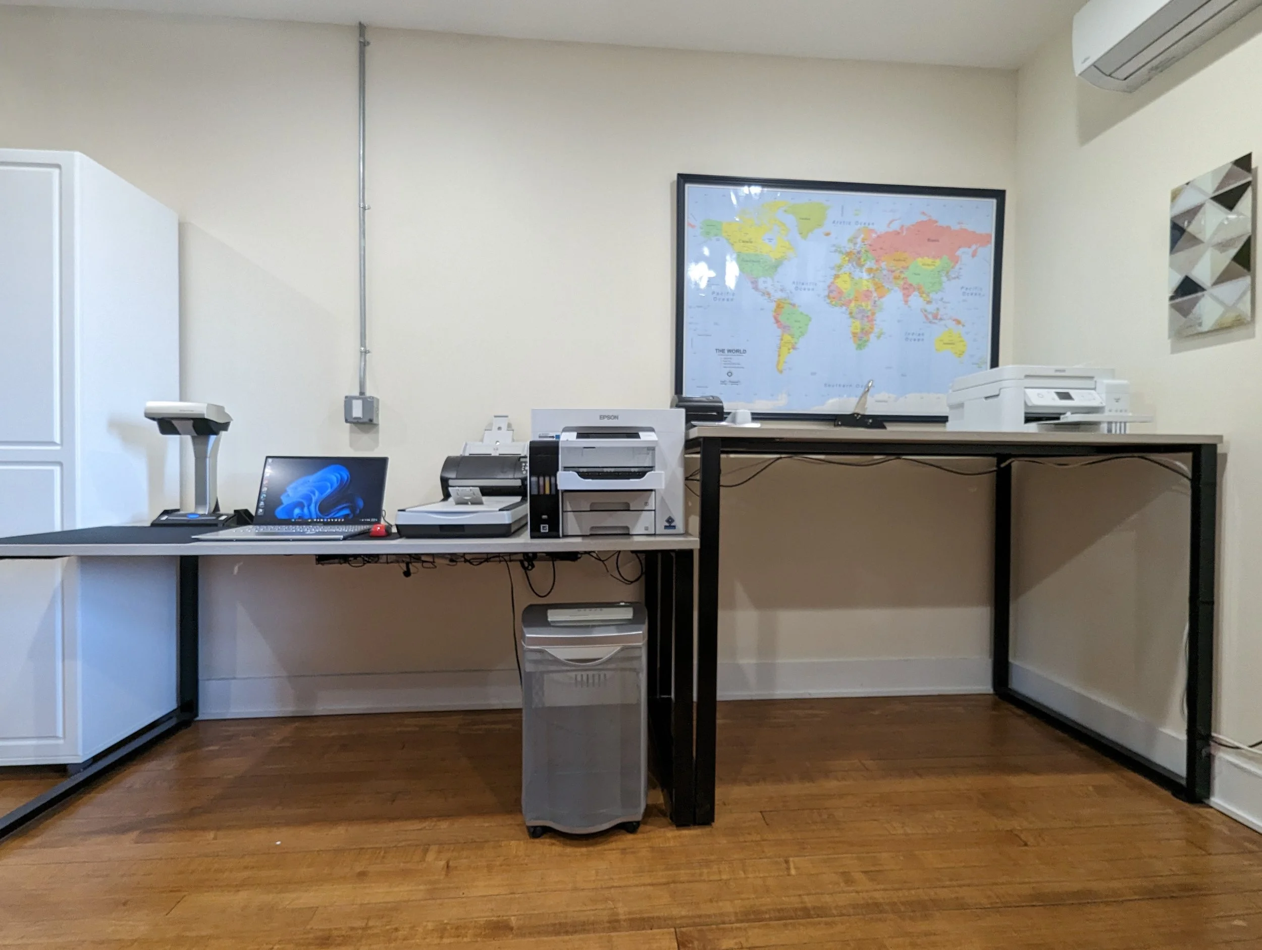 Office workspace with a desk holding a laptop, printers, and a scanner, against a wall with a world map and abstract artwork, and a cabinet on the left.