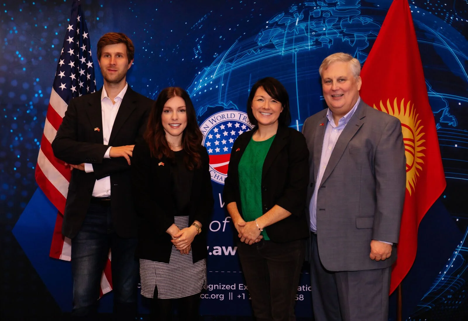 Group of five people standing in front of a backdrop with American and Kyrgyz flags, and a logo, at an official event.