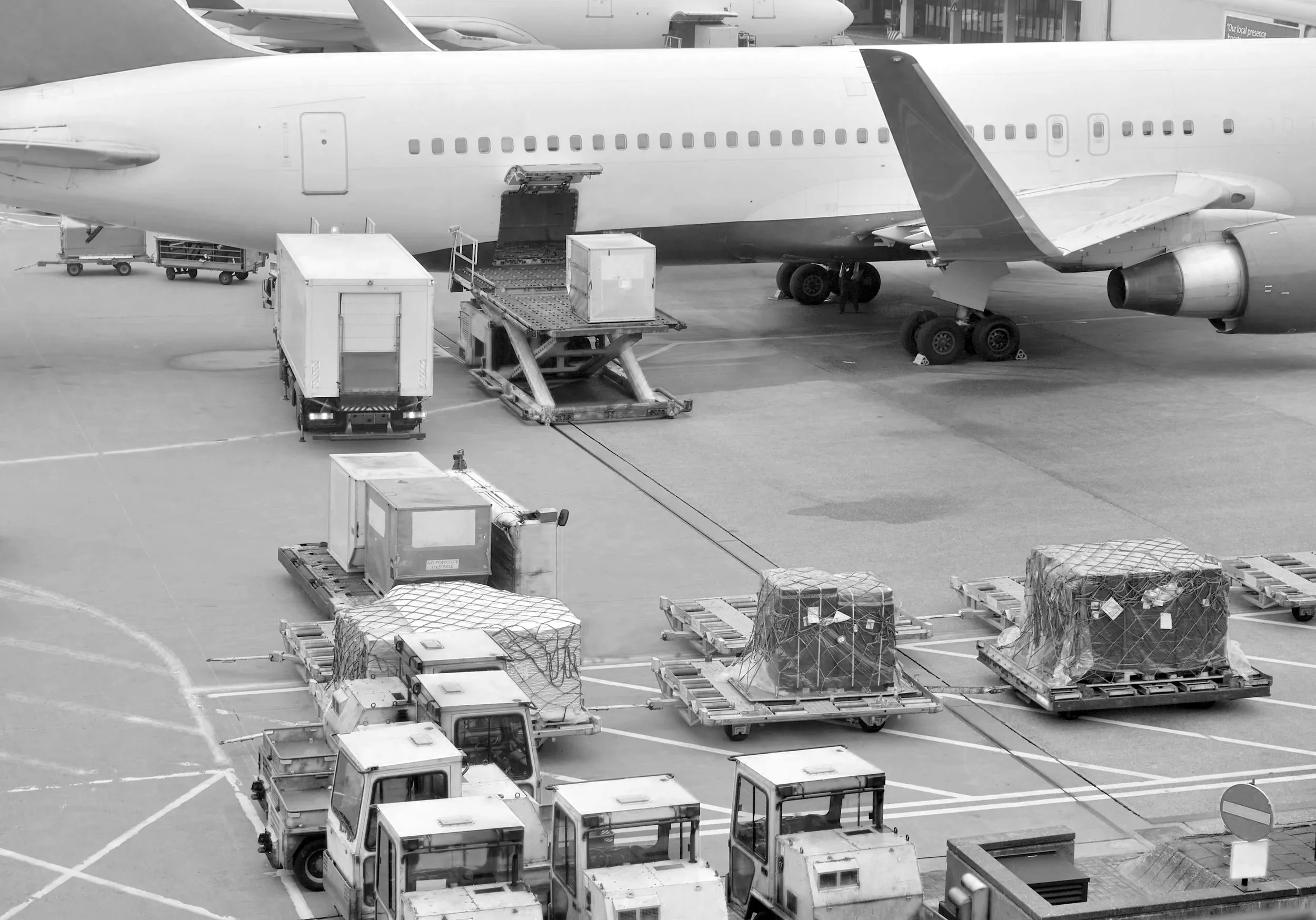 An airplane parked at an airport gate with cargo being loaded or unloaded using various ground service vehicles and equipment.