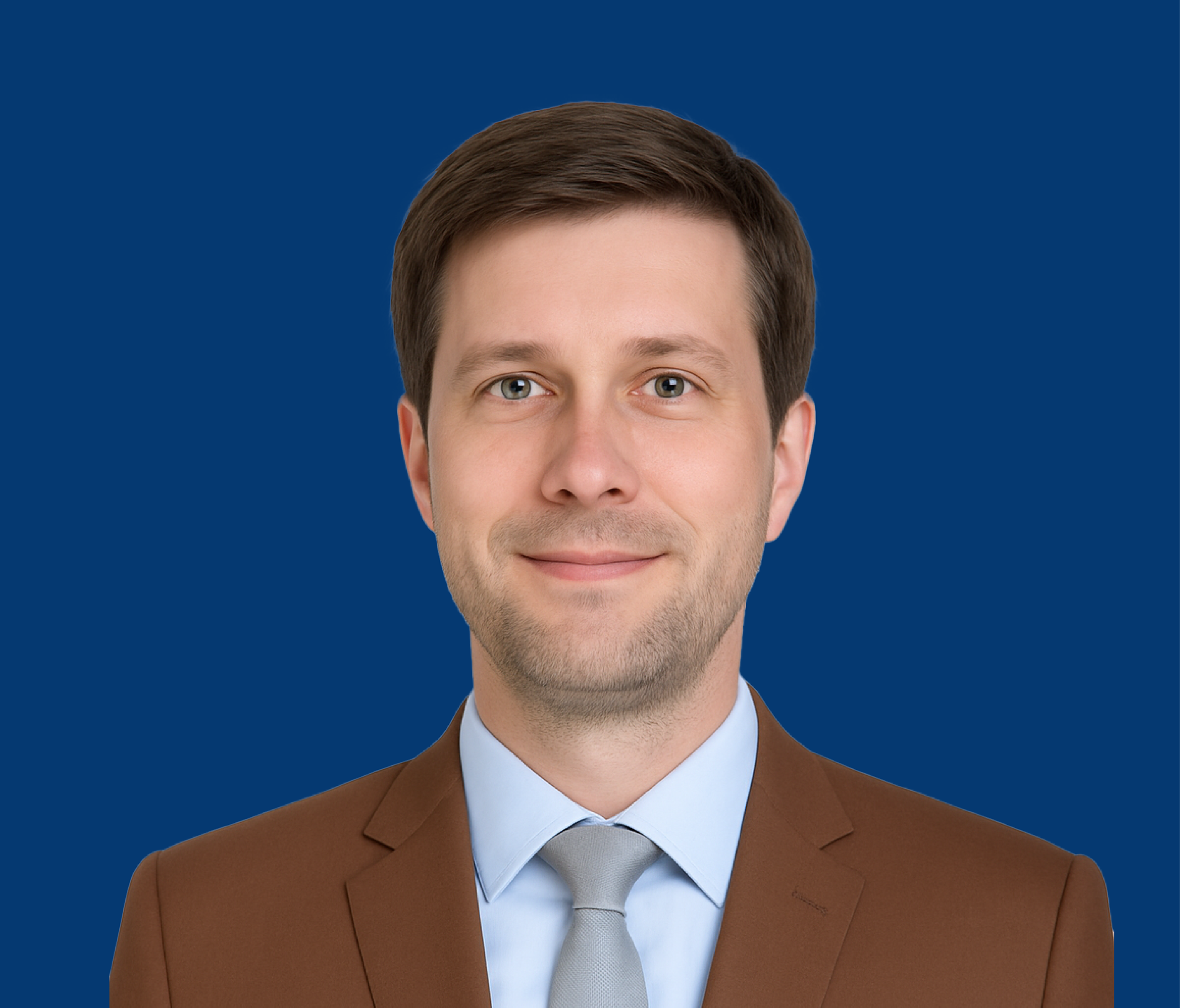A professional man with brown hair, wearing a brown suit, blue shirt, and light gray tie, smiling against a solid blue background.