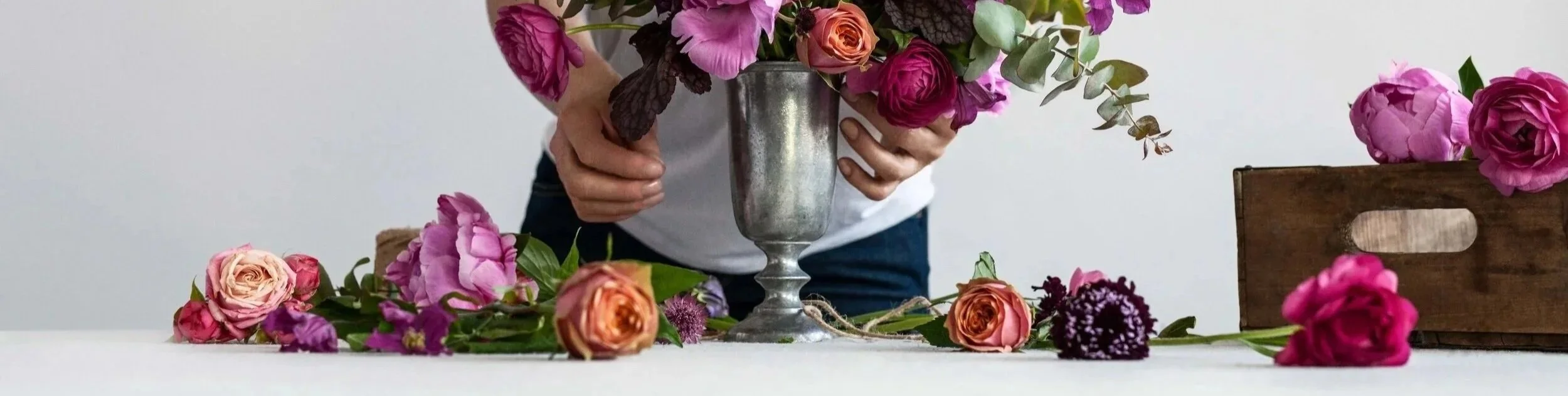 A person arranging pink and orange roses and purple flowers in a silver vase on a white table, with additional flowers and a wooden box nearby.