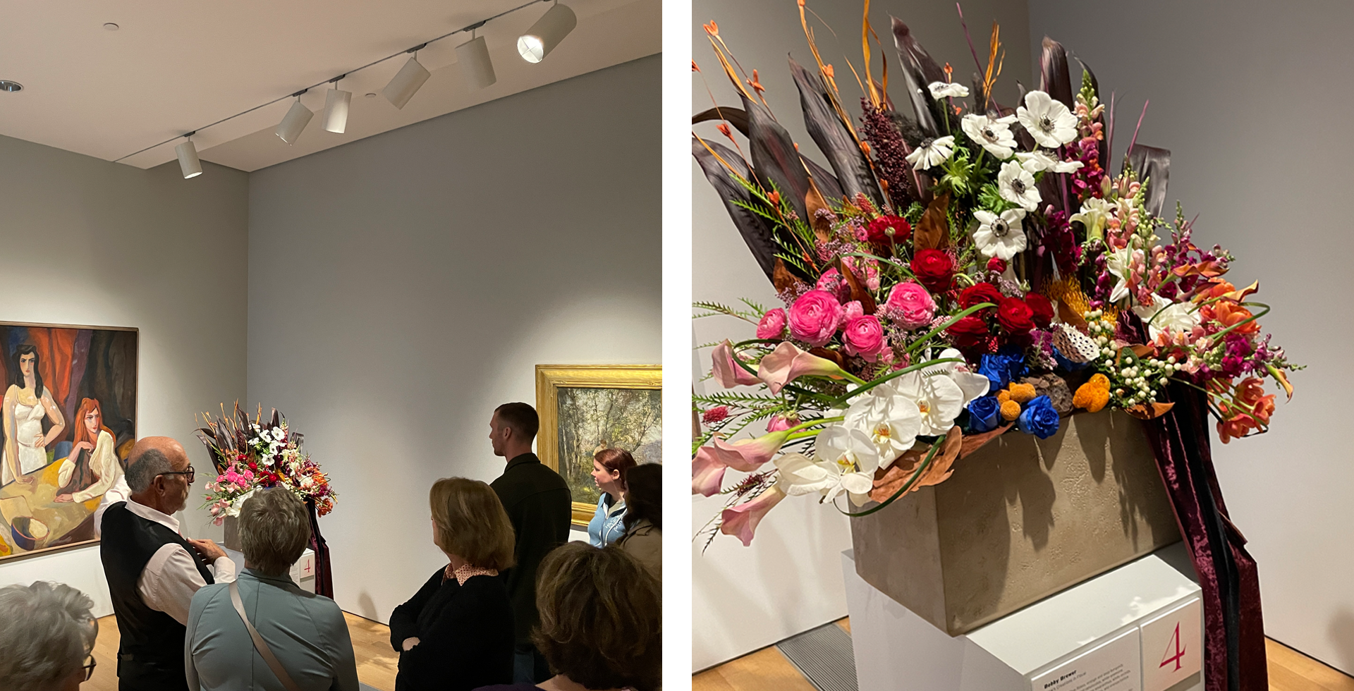 People viewing a colorful flower arrangement at an art gallery, where paintings are displayed on the walls and spotlights illuminate the artwork and floral display.