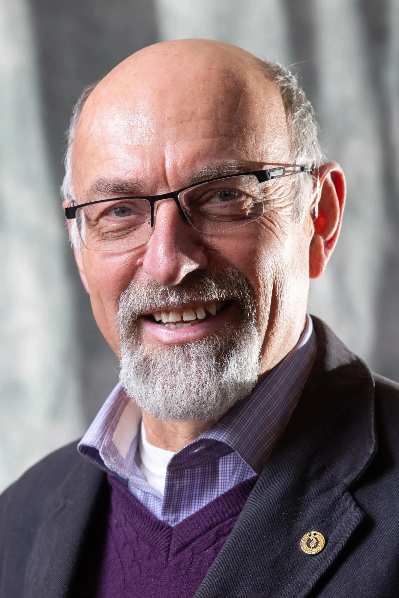 Close-up of a smiling older man with glasses, a bald head, grey beard, wearing a dark blazer, purple sweater, and collared shirt against a blurred background.