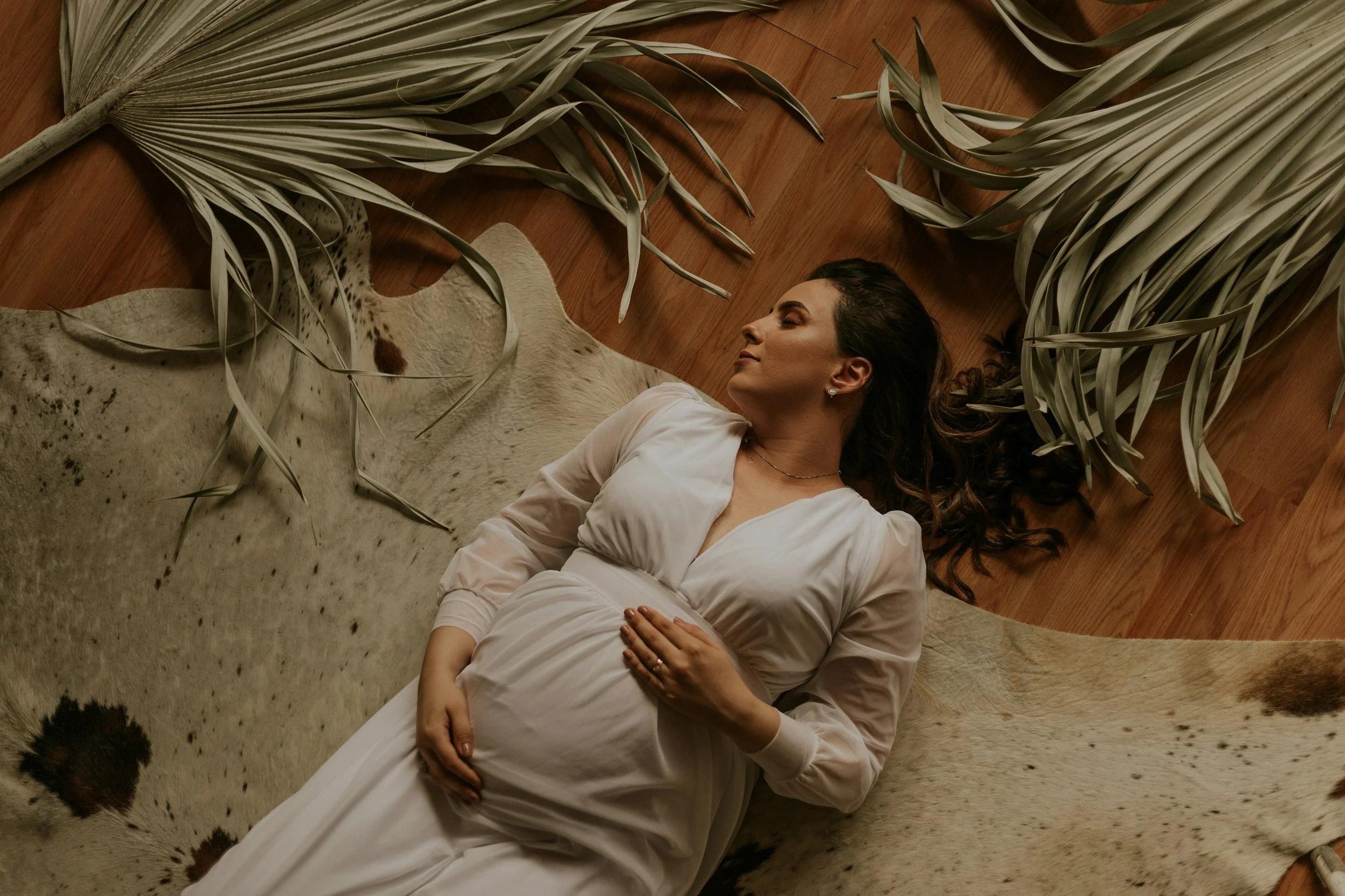 A pregnant woman in a white dress lying on a cowhide rug surrounded by large dried palm leaves.