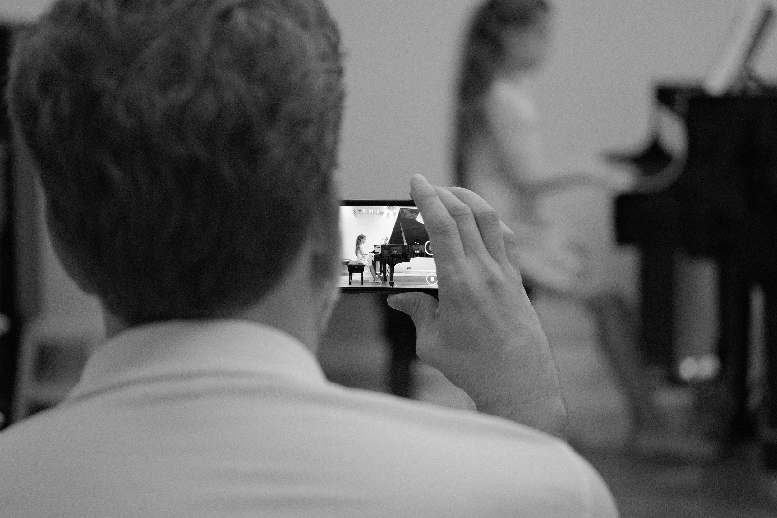 A person taking a photo of a girl at a grand piano on a smartphone in a music rehearsal room.