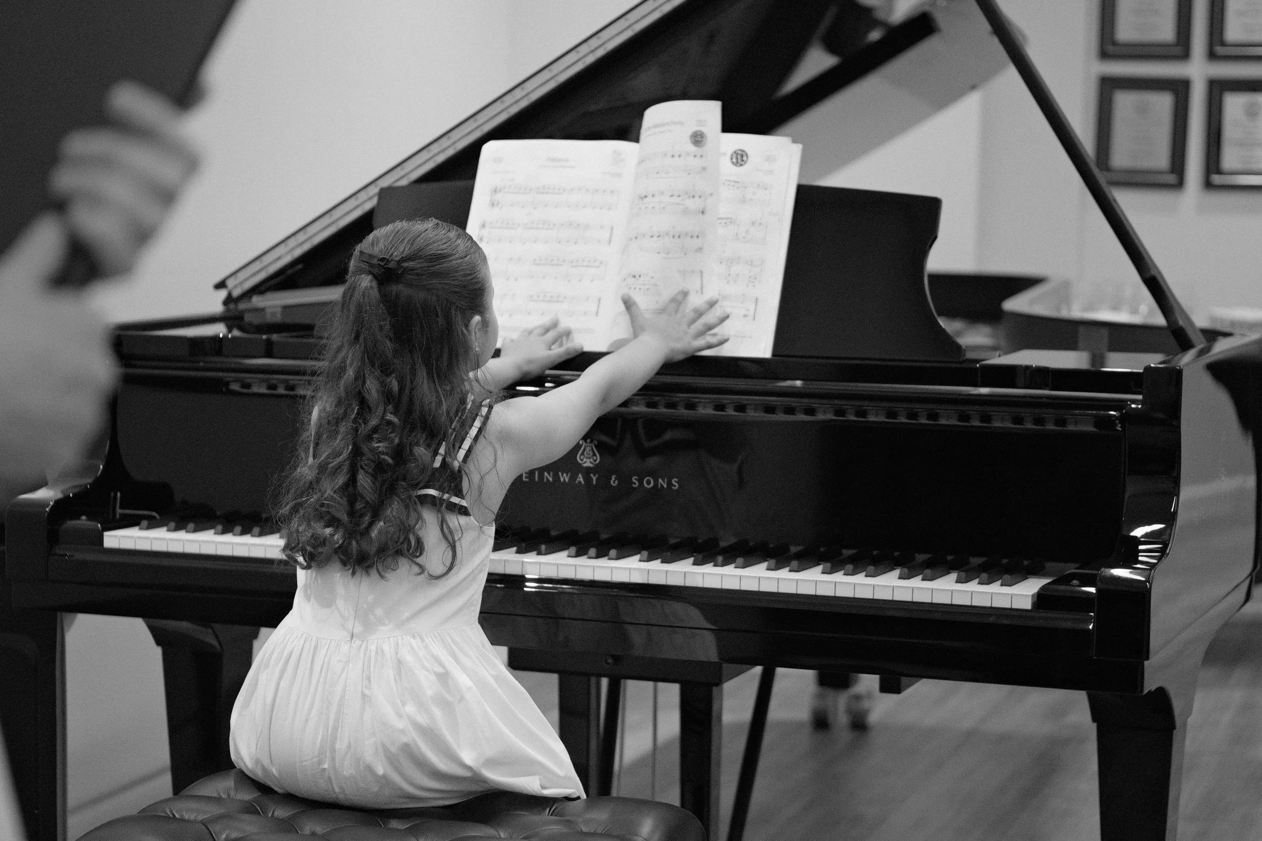 A young girl with long curly hair in a dress playing a grand piano with sheet music in front of her.