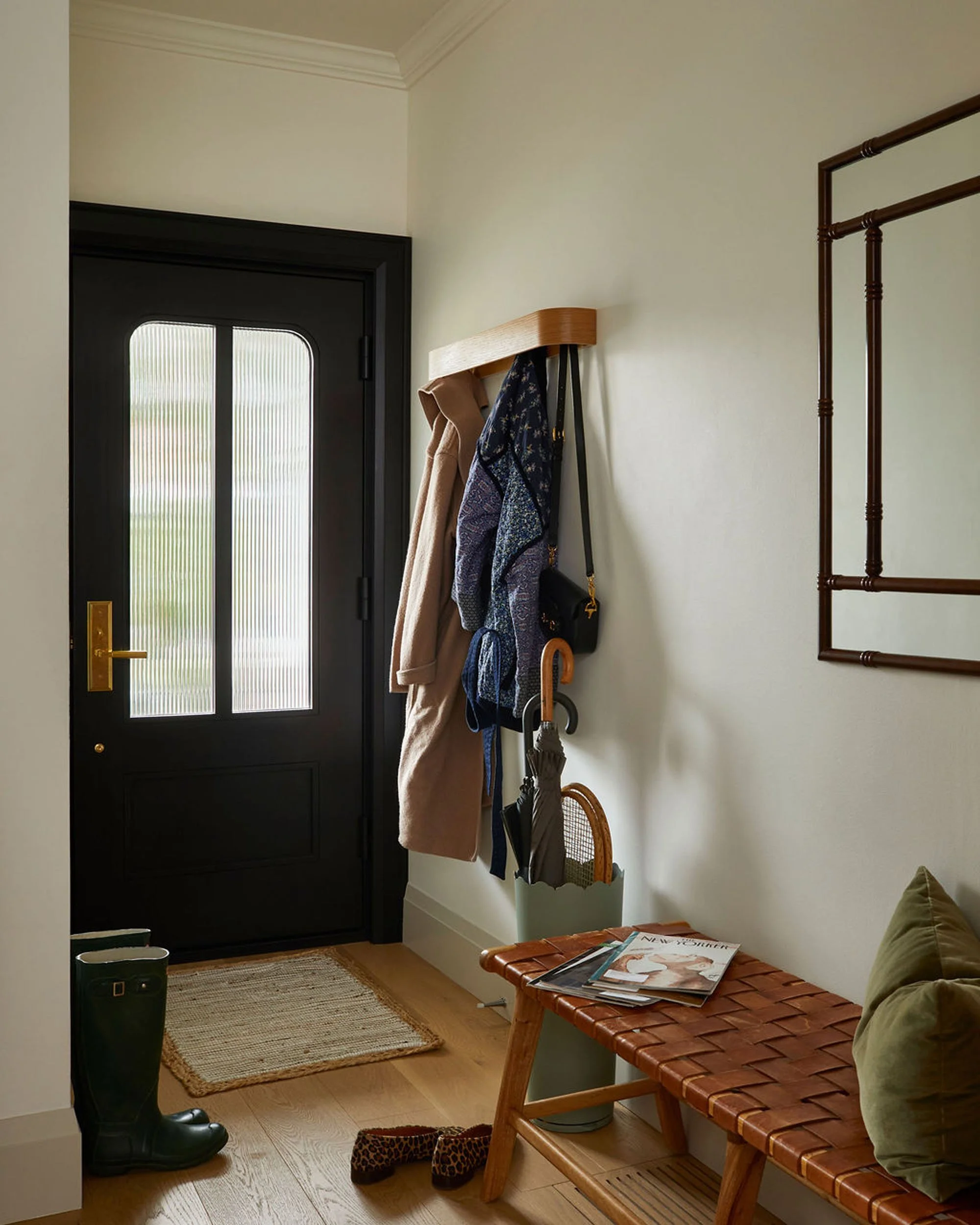 Entryway with a black front door, a coat and accessories hanging on a wall-mounted wooden rack, a pair of black rain boots, a small woven rug, and a brown leather bench with magazines on top.