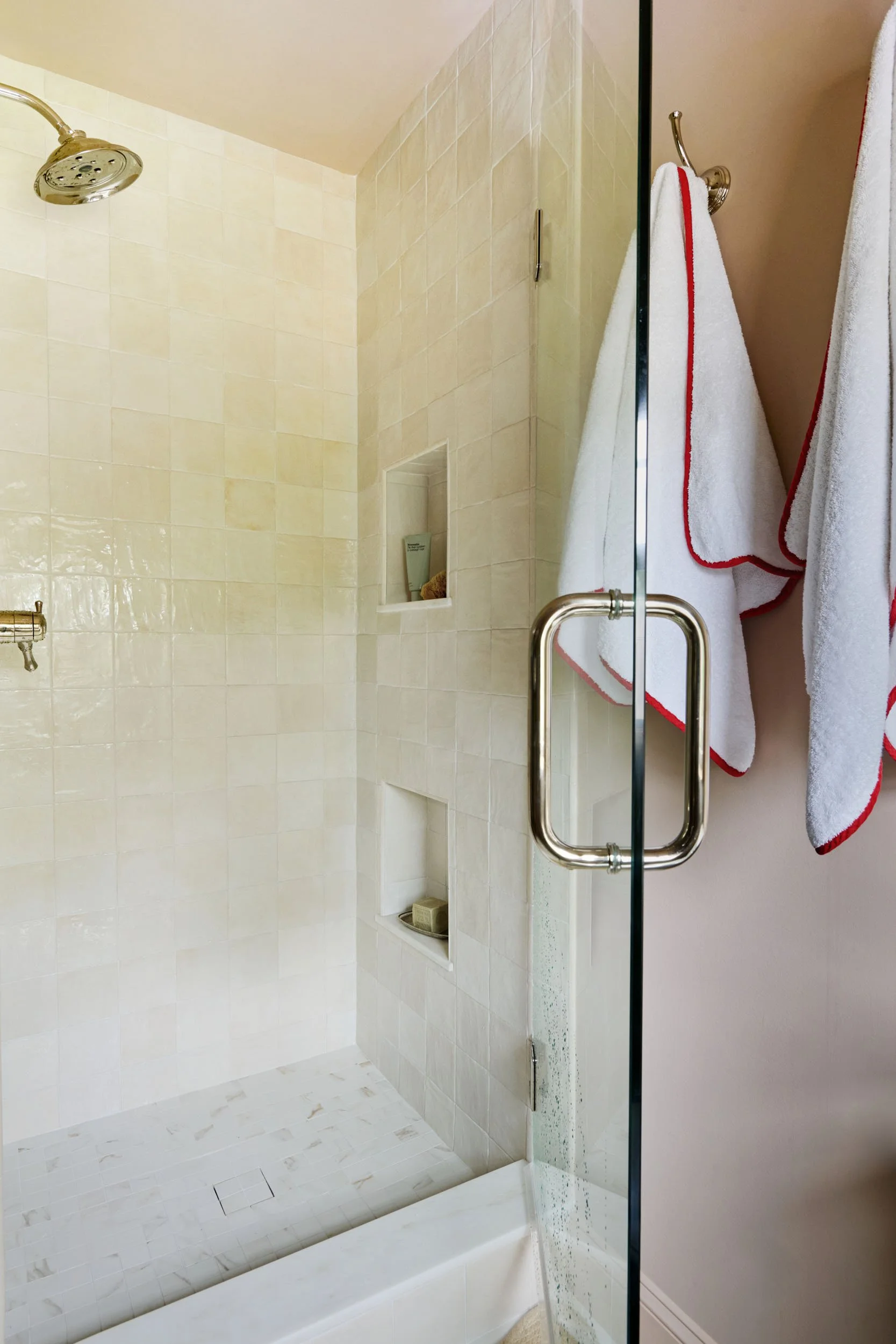 Shower with beige tile walls, a rainfall showerhead, built-in niche with shampoo and soap, and two white towels with red trim hanging on hooks outside the glass door.