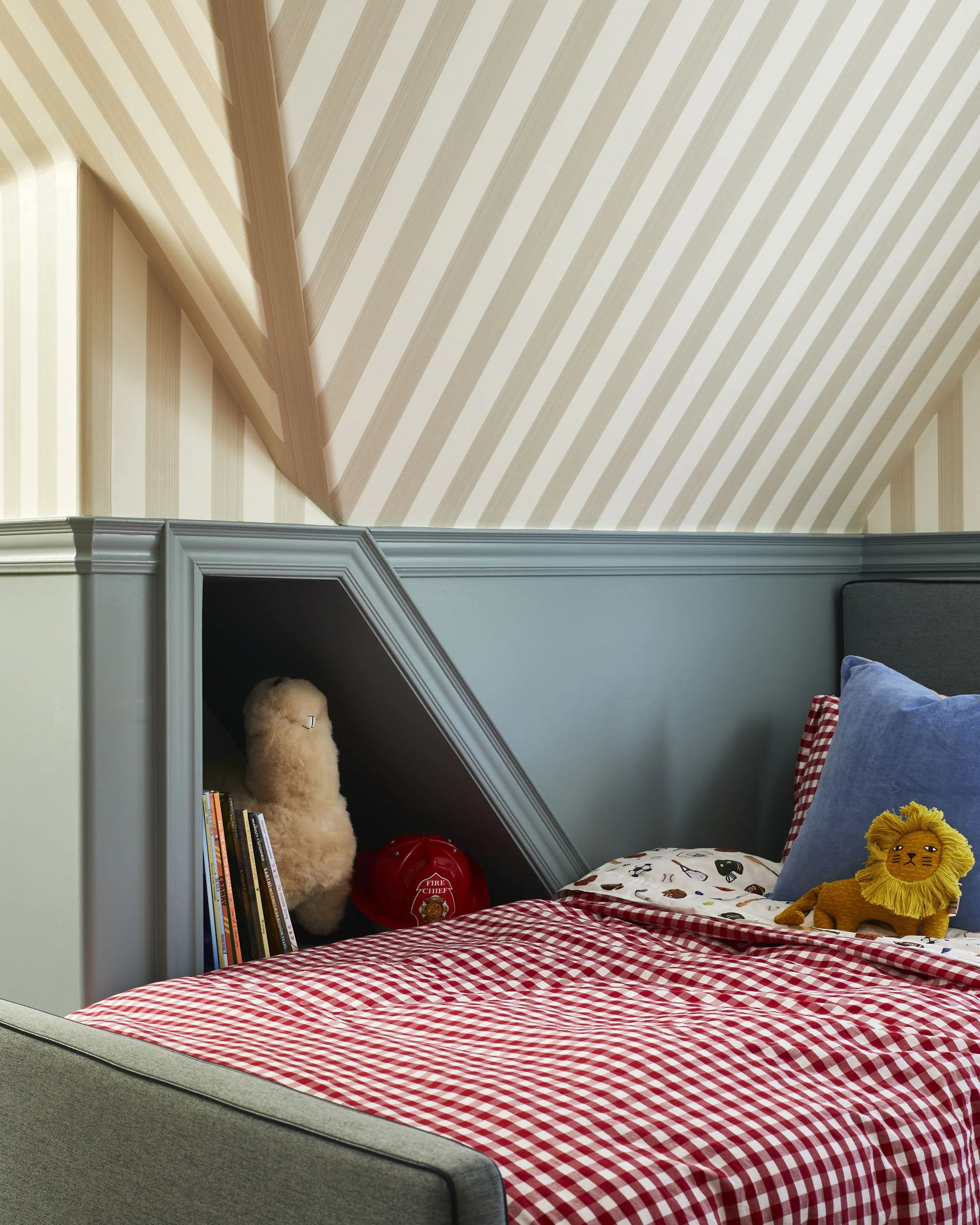 A cozy bedroom with a bed covered in a red and white checkered blanket, blue and red pillows, and a stuffed lion toy. The room has a sloped ceiling with light wood paneling and a built-in shelf containing books, a plush llama, a red fire alarm, and a