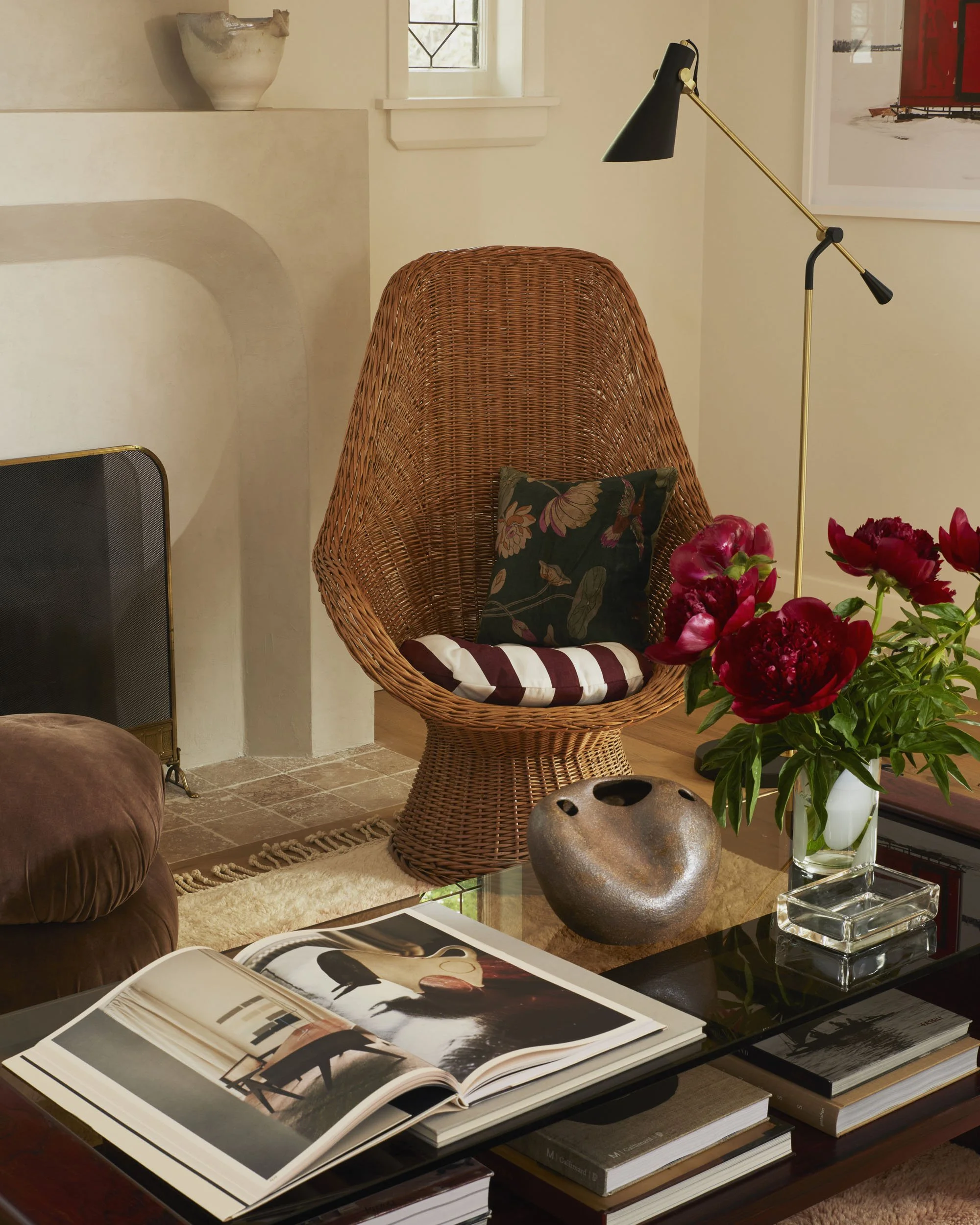 A wicker accent chair with cushions and a pillow, next to a glass coffee table with an open magazine, a decorative ceramic piece, a vase of peonies, and books in a cozy living room.