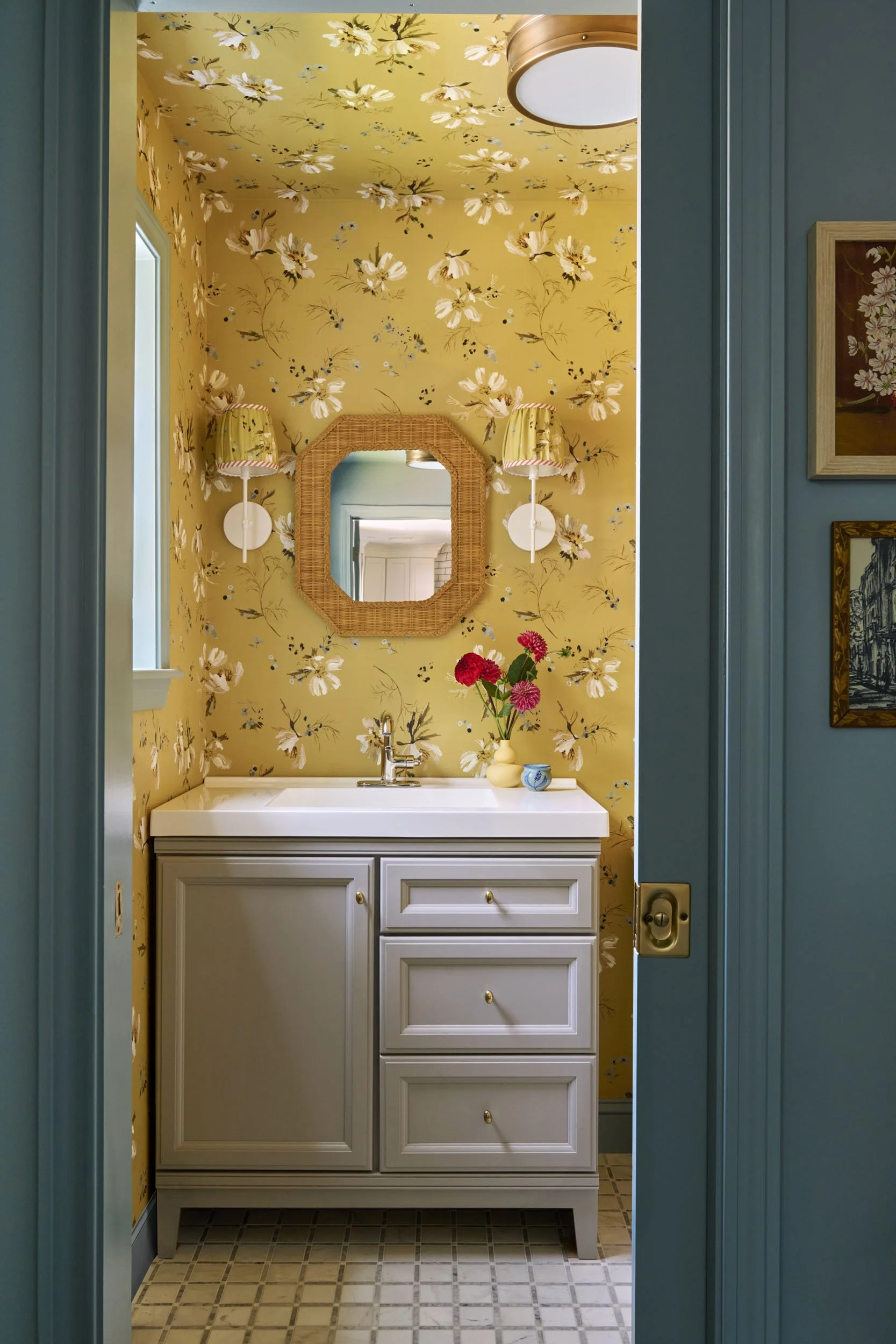 A small bathroom with yellow floral wallpaper, a white vanity with gold knobs, a mirror with a rattan frame, and wall sconces with yellow patterned shades. There are pink and red flowers in a cream vase on the vanity.