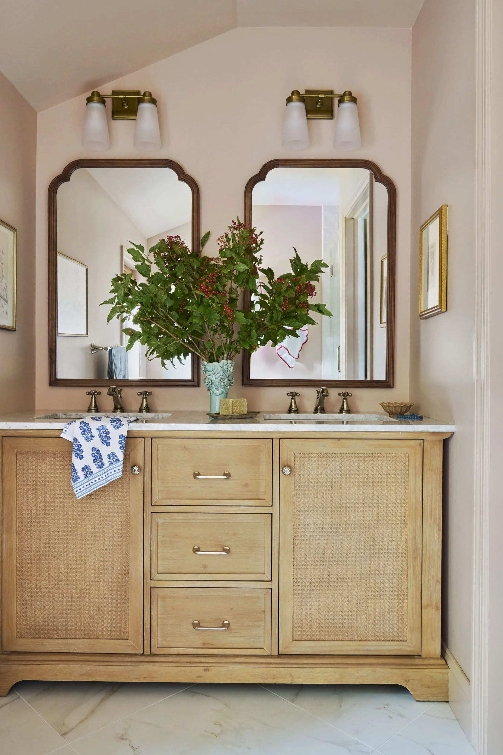Bathroom vanity with two mirrors, a large floral arrangement in a light blue vase, gold fixtures, a soap dish, and a towel hanging over a drawer handle.