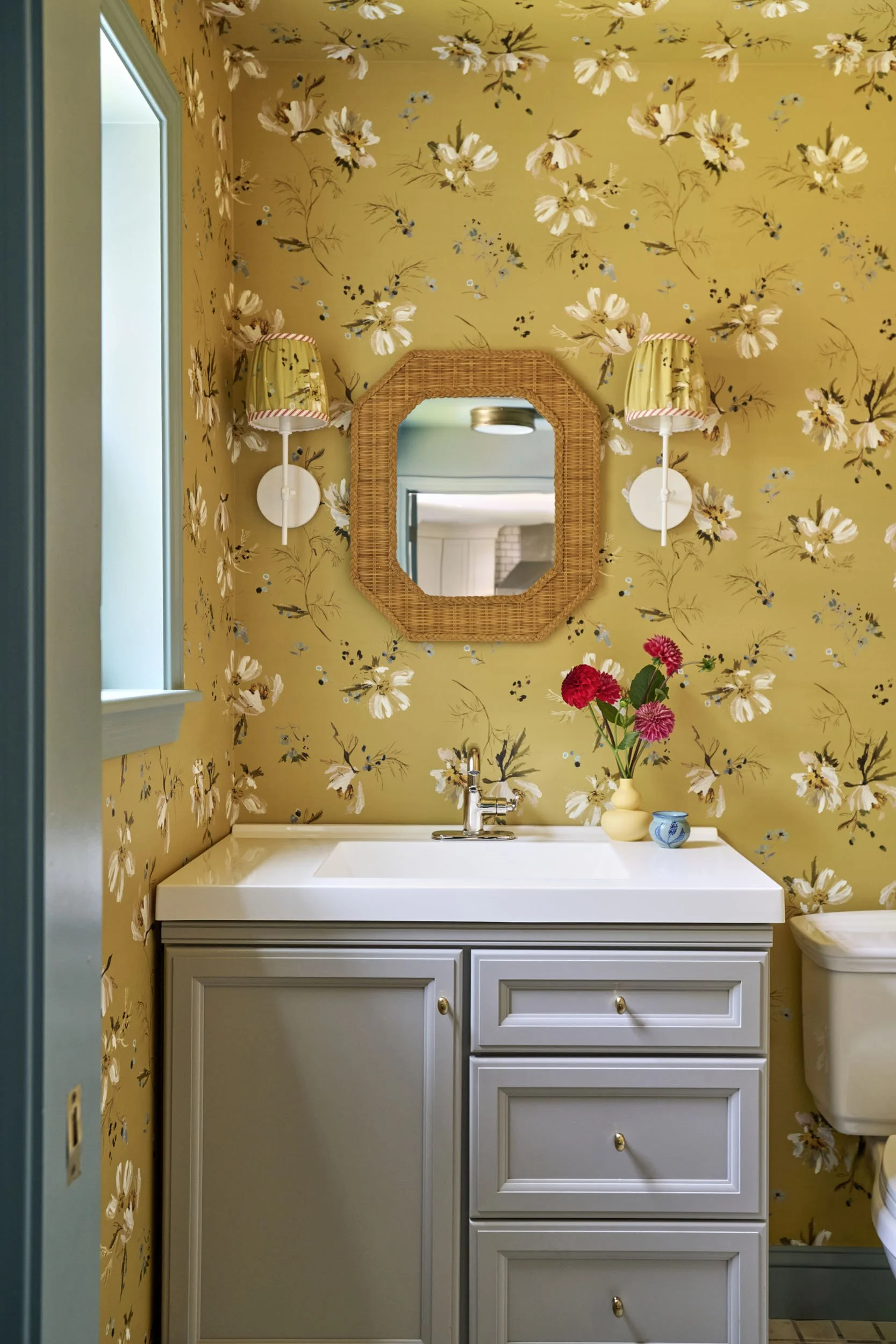 A small bathroom with yellow floral wallpaper, a white sink cabinet, a mirror with a wicker frame, two wall-mounted lamps with yellow and floral lampshades, a window with blue walls, and a vase of pink flowers on the sink.