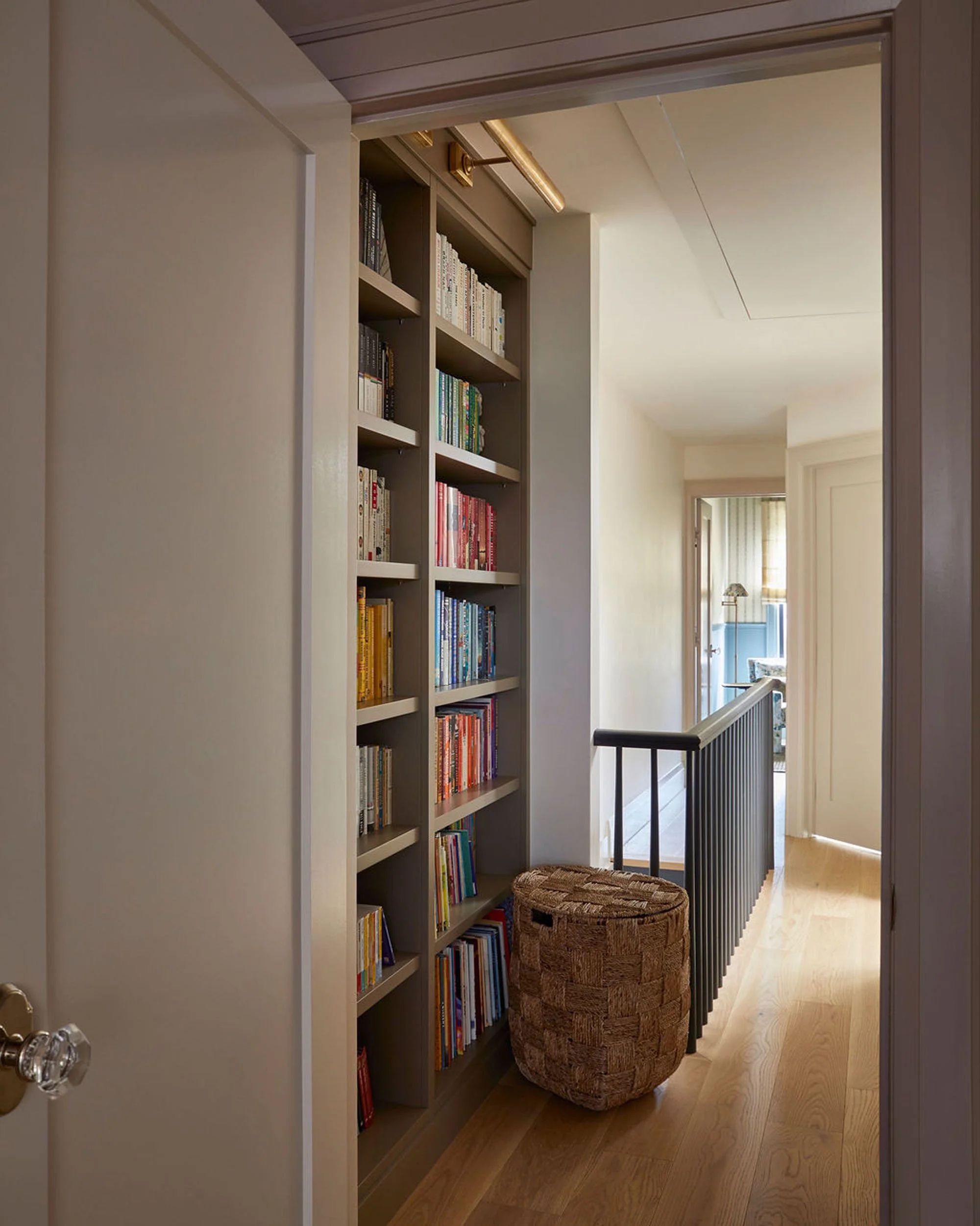 Hallway with a built-in bookshelf filled with books, a woven ottoman, and a staircase with a black handrail leading to a sunlit room.