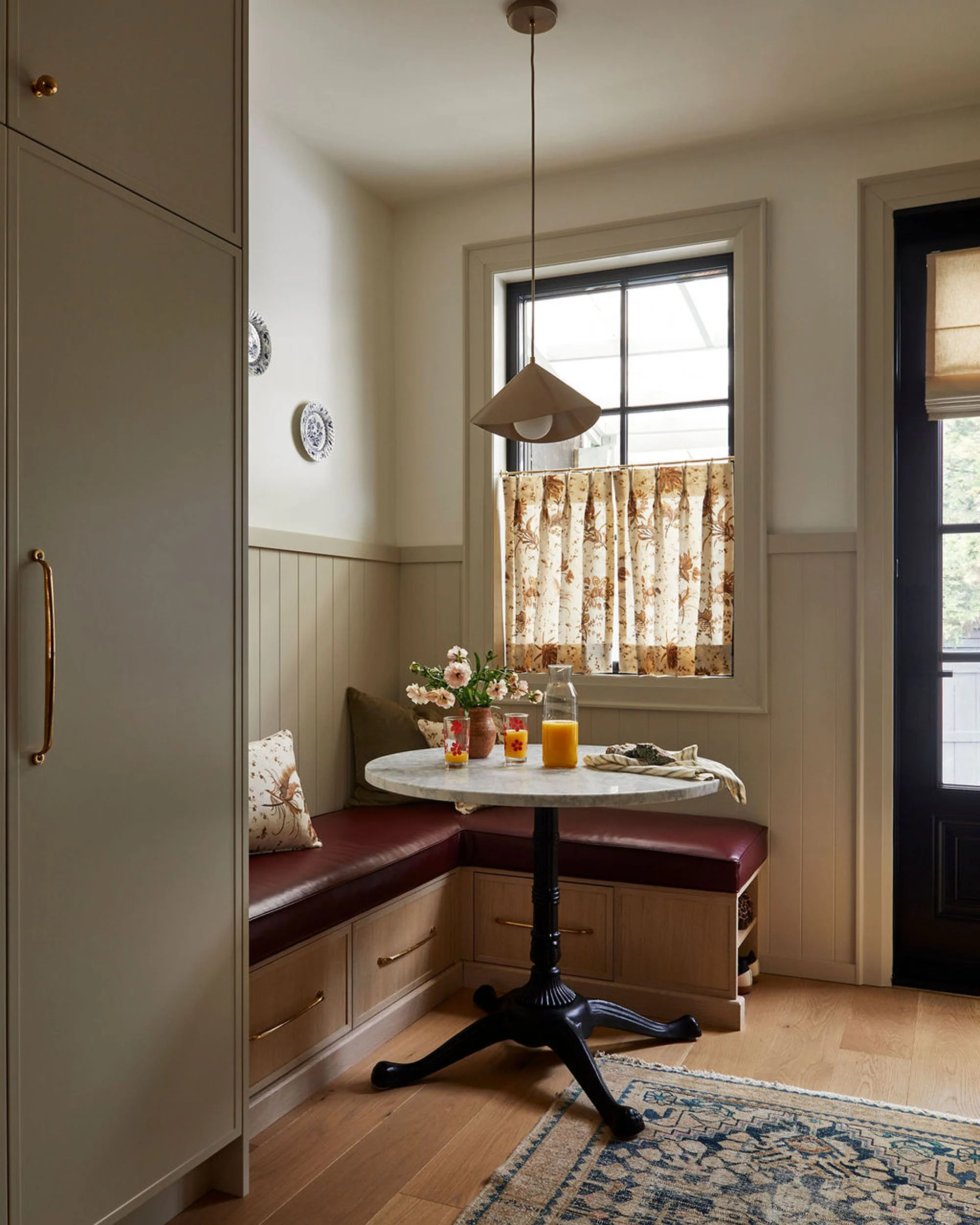 A cozy corner of a kitchen with a built-in bench seat, a small round marble table, a vase with pink flowers, glasses of orange juice, and window with floral curtains.
