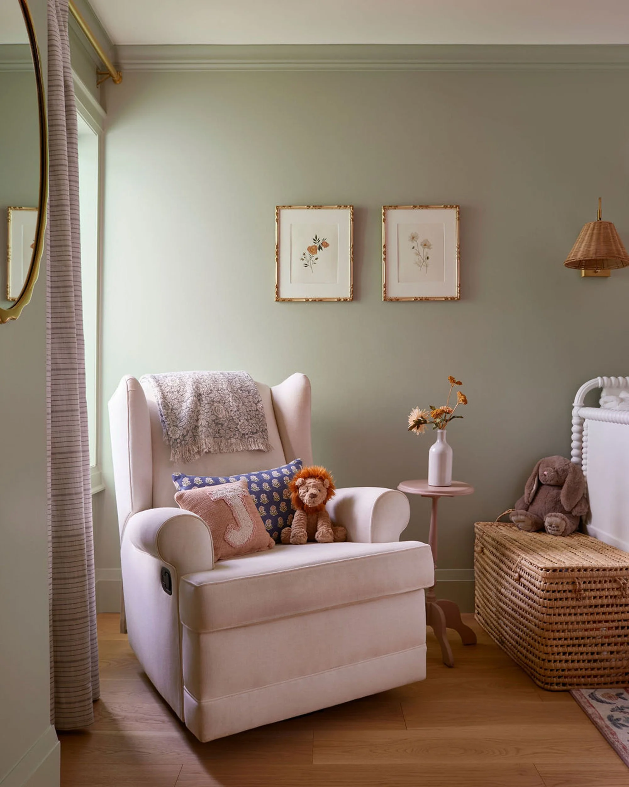 A cozy nursery corner featuring a white armchair with a gray throw blanket and decorative pillows, including a pink one with a letter J, and a stuffed lion. Beside the chair is a pink side table with a white vase and flowers. To the right is a wicker
