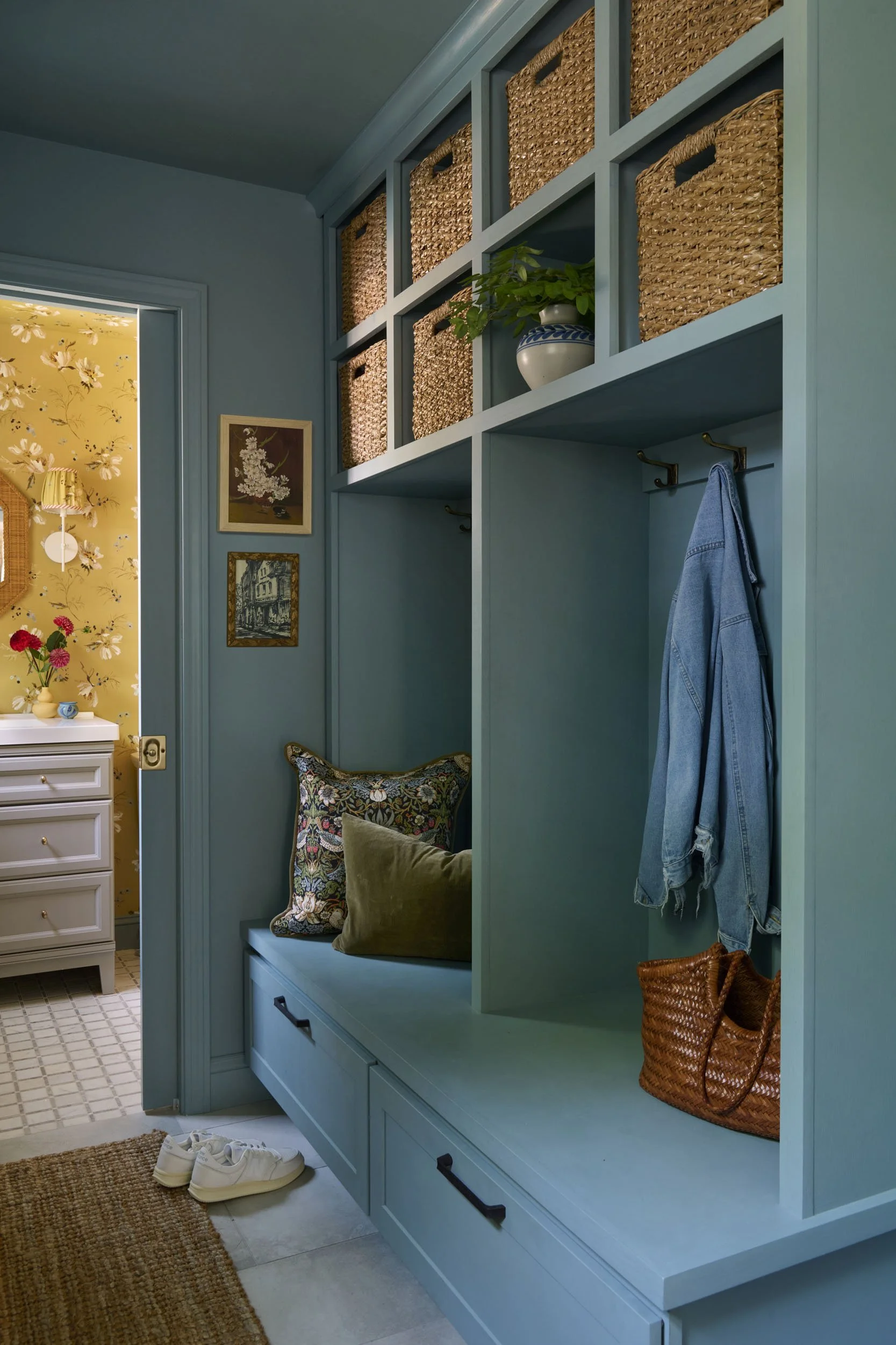 Interior view of a blue built-in storage bench with cushions, hanging hooks with a denim jacket, woven baskets on the upper shelves, and a pair of white sneakers on the floor. Adjacent room with yellow floral wallpaper and white furniture is visible 