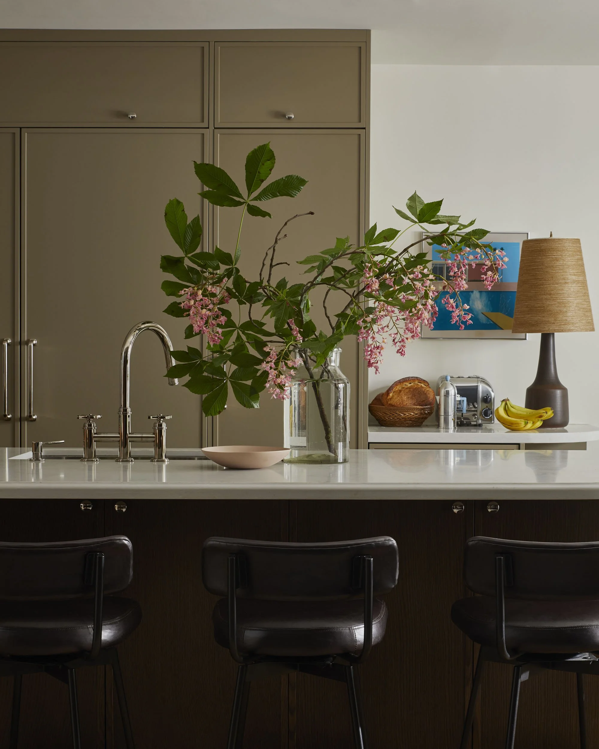 Kitchen island with a large glass vase holding pink flowering branches, a pink bowl, and three dark brown barstools.