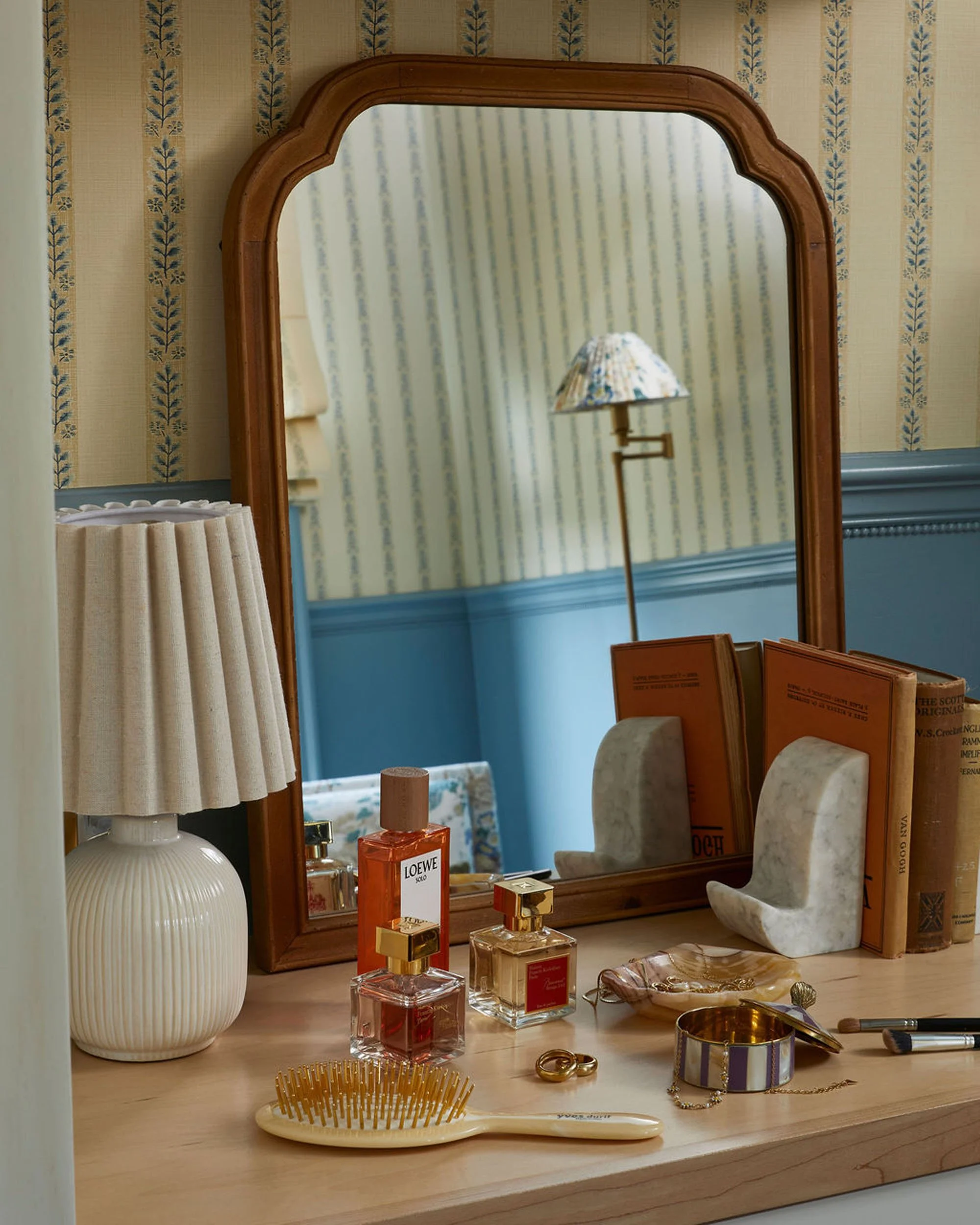 A wooden dresser with a mirror reflecting a striped wallpapered wall and a floor lamp with a floral lampshade. Various perfume bottles, jewelry, a hairbrush, and decorative items are arranged on the dresser.