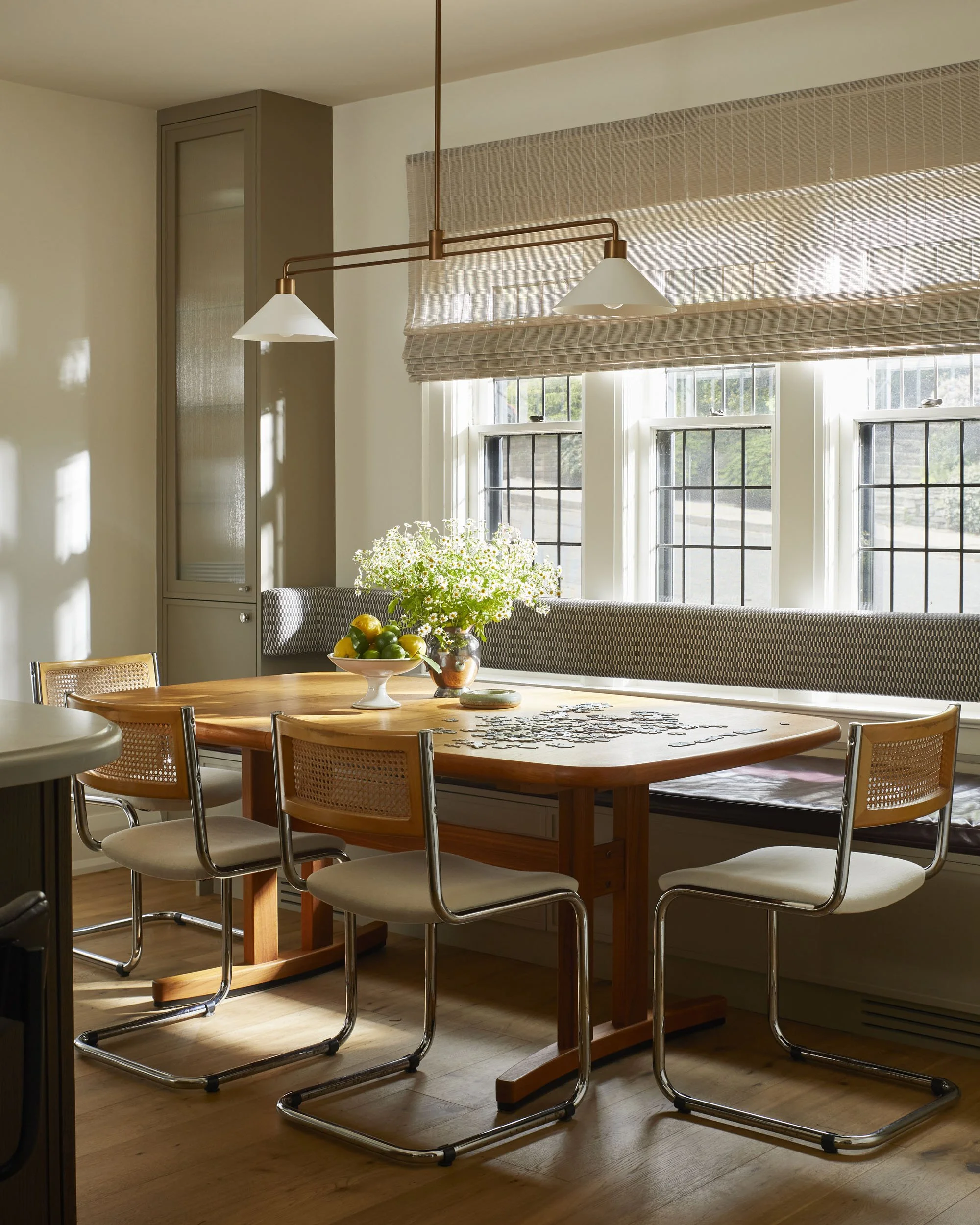 Dining room with wooden table, four modern chairs, window bench, and large windows with blinds, decorated with a vase of flowers and a bowl of fruit, natural sunlight
