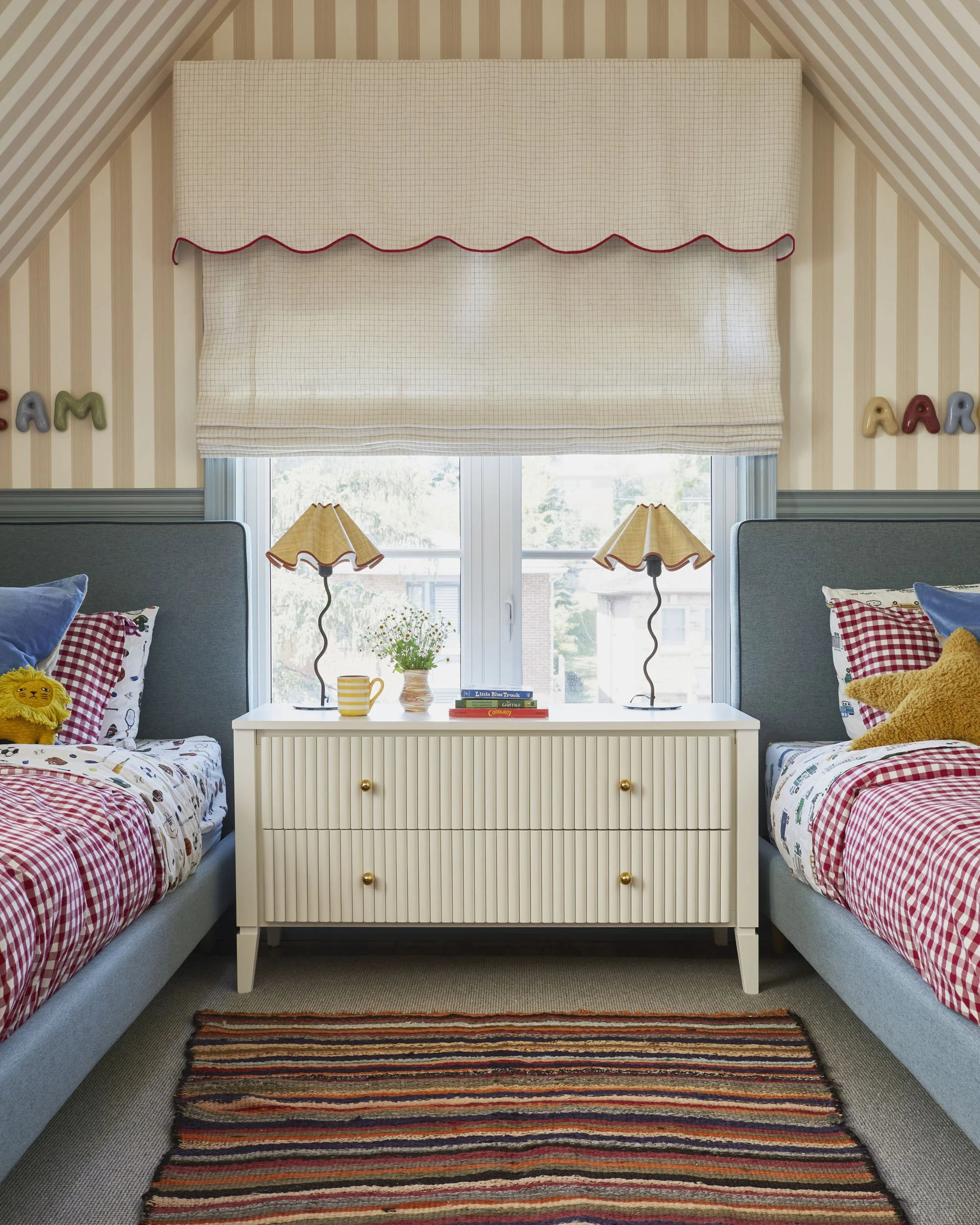 A cozy bedroom with two beds covered in red and white checkered bedding, teddy bears and colorful pillows, a white dresser in between with two yellow lamps, a window with a beige valance, and striped wallpaper.