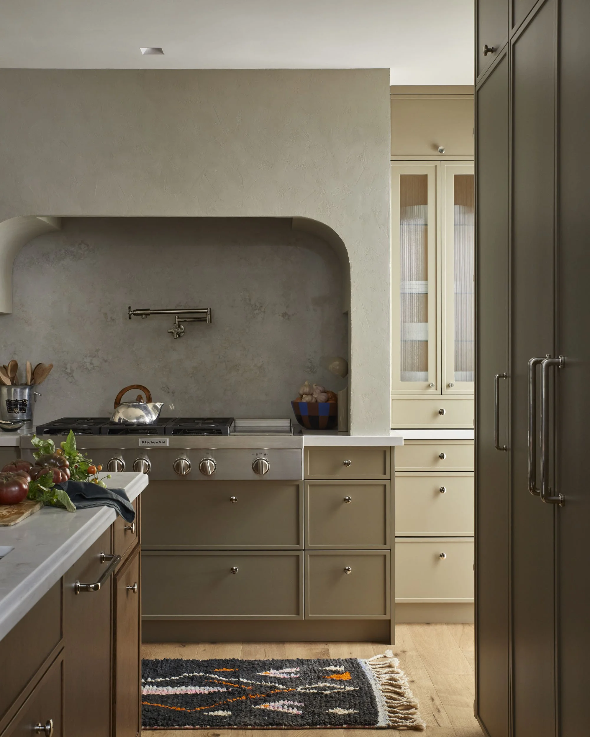 A modern kitchen with beige and dark wood cabinetry, stainless steel appliances, a textured wall, a colorful rug on wooden flooring, and various kitchen utensils and ingredients on the counters.