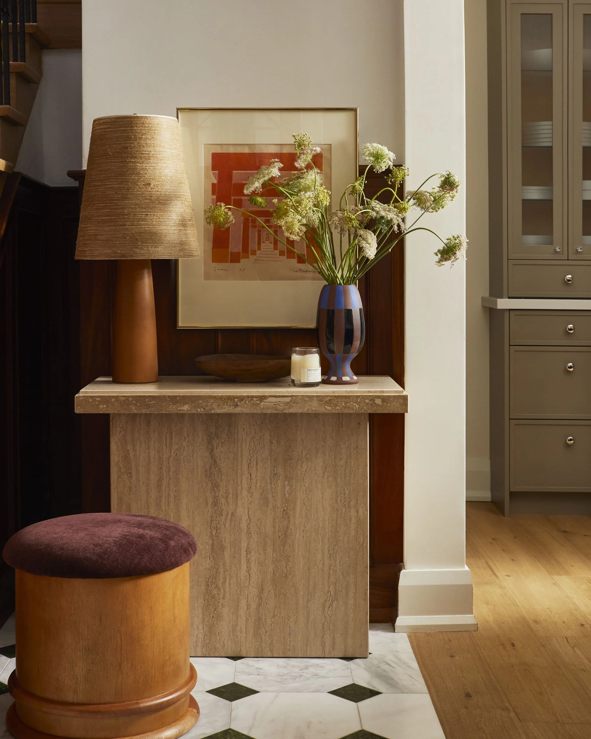 A decorative console table with a brown table lamp, a framed abstract artwork, a bowl, a candle, and a vase with white flowers, located in a room with wood and tiled flooring.