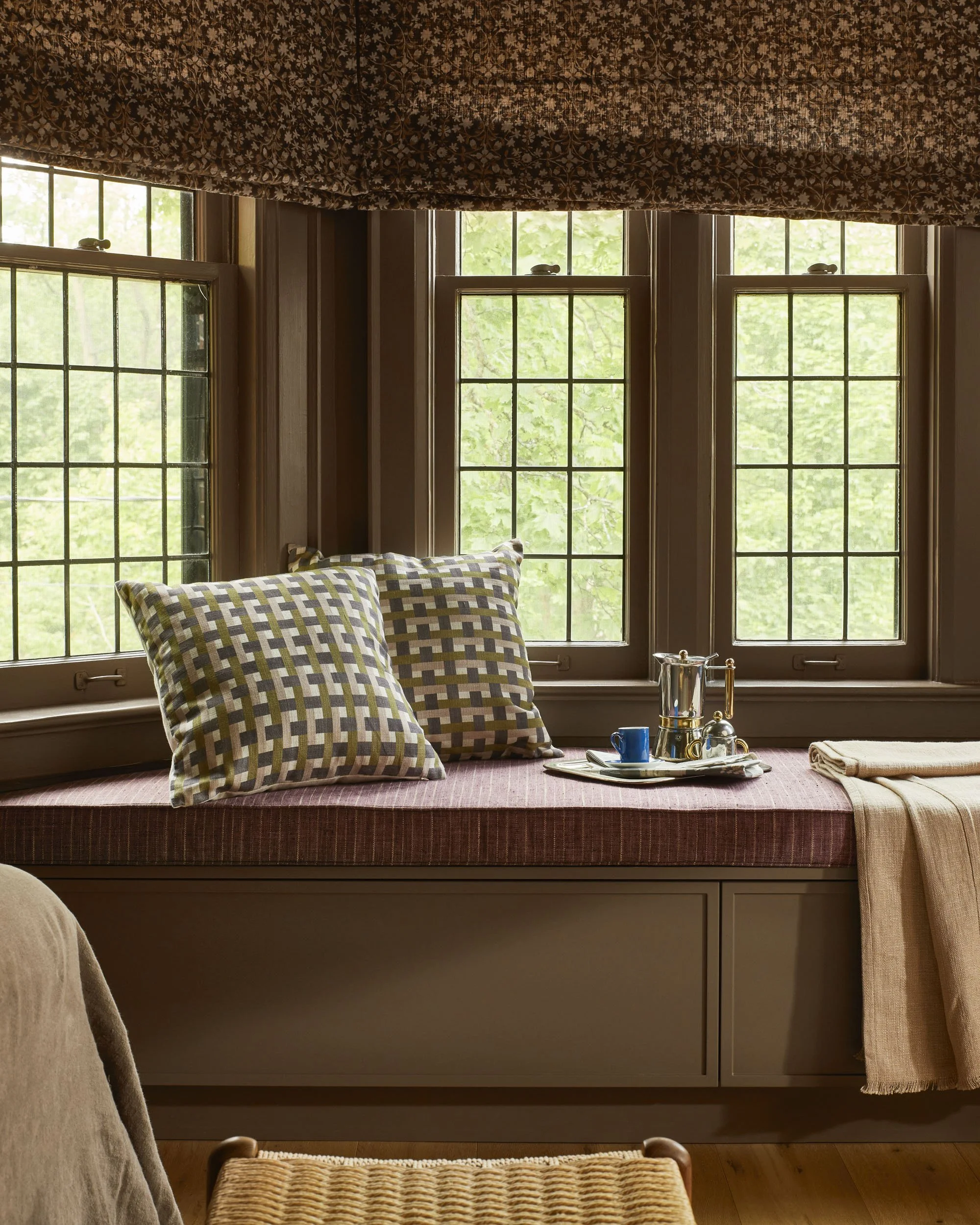 A window seat with two patterned pillows, a tea set on a tray, and a beige blanket, in front of three large windows with lush green trees outside, under a brown patterned valance.