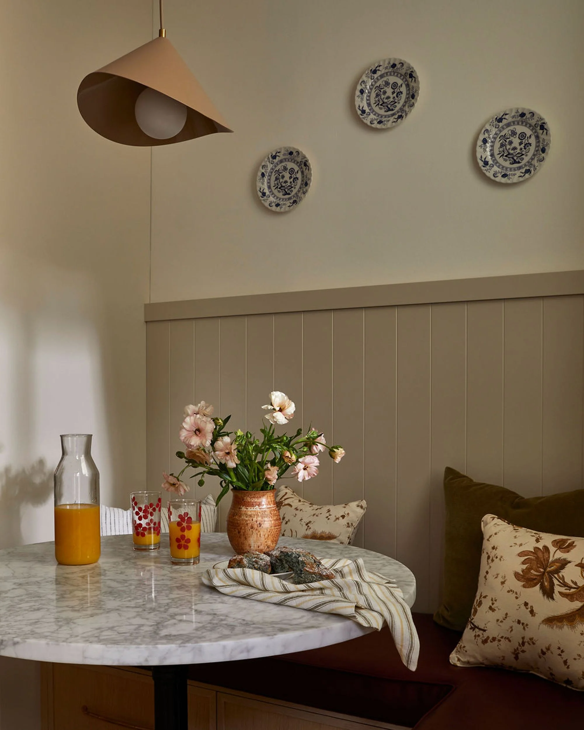 A dining area with a marble table featuring a vase of pink flowers, a glass pitcher, two glasses of orange juice, and some rocks on a striped cloth. There is a cushioned bench with floral pillows and three decorative plates on the wall. A modern beig