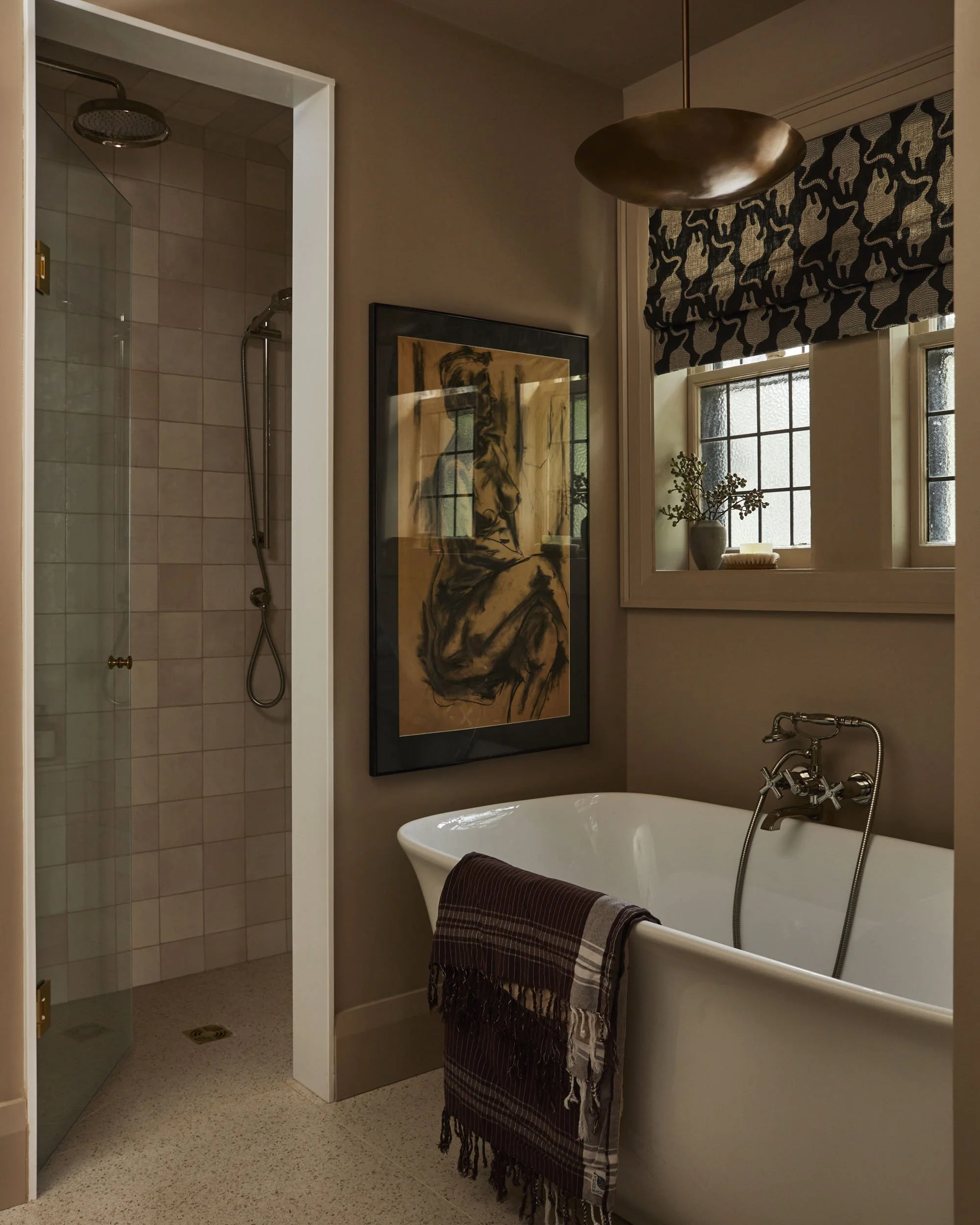 A bathroom with a clawfoot bathtub, a window with black and white patterned Roman shade, a potted plant, a framed artwork of a seated woman, and a walk-in shower with glass door.