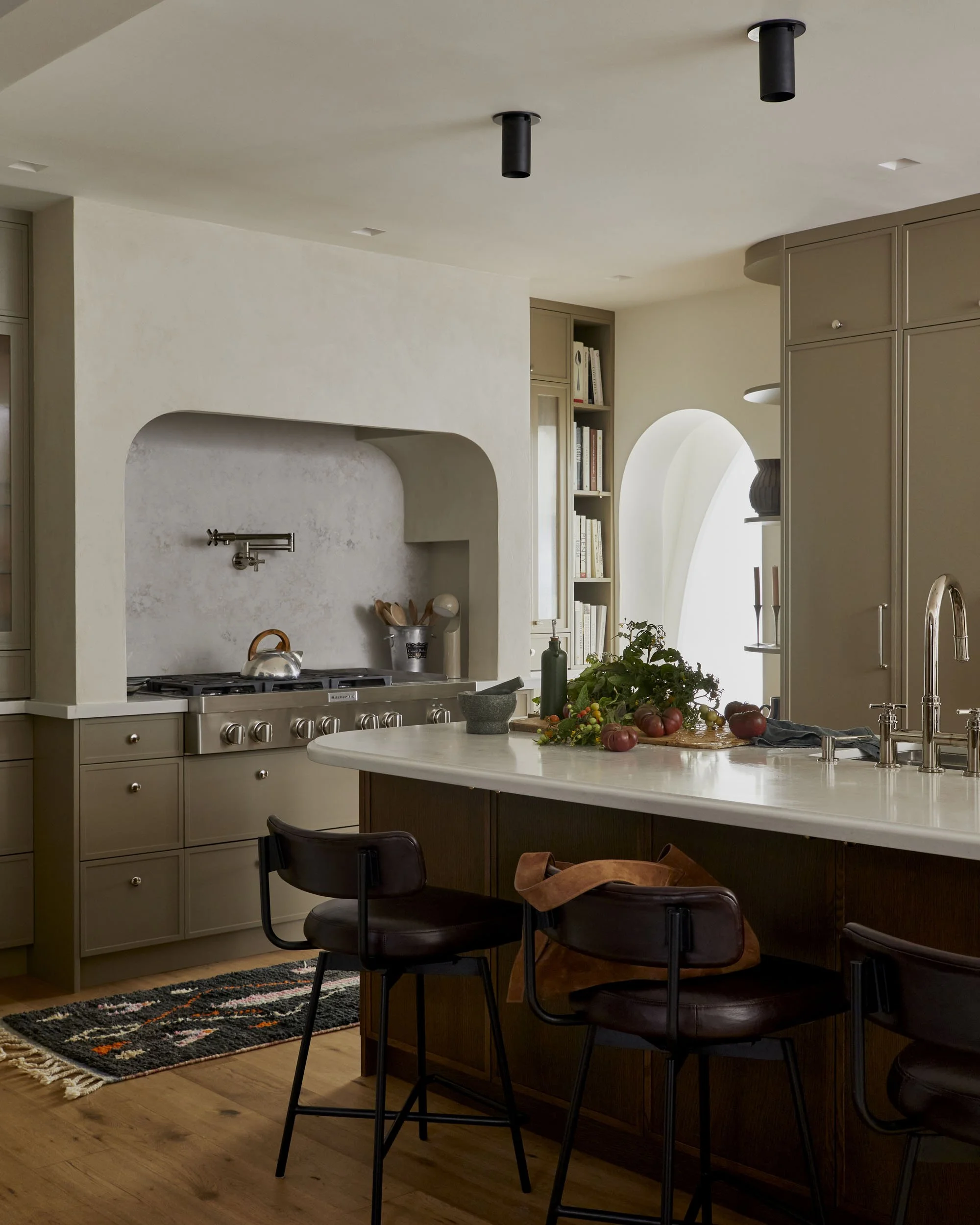 A modern kitchen with beige cabinetry, a white curved wall, a kitchen island with a marble countertop, and black bar stools. There are bookshelves, a window with natural light, and a countertop with fresh vegetables and a mortar and pestle.
