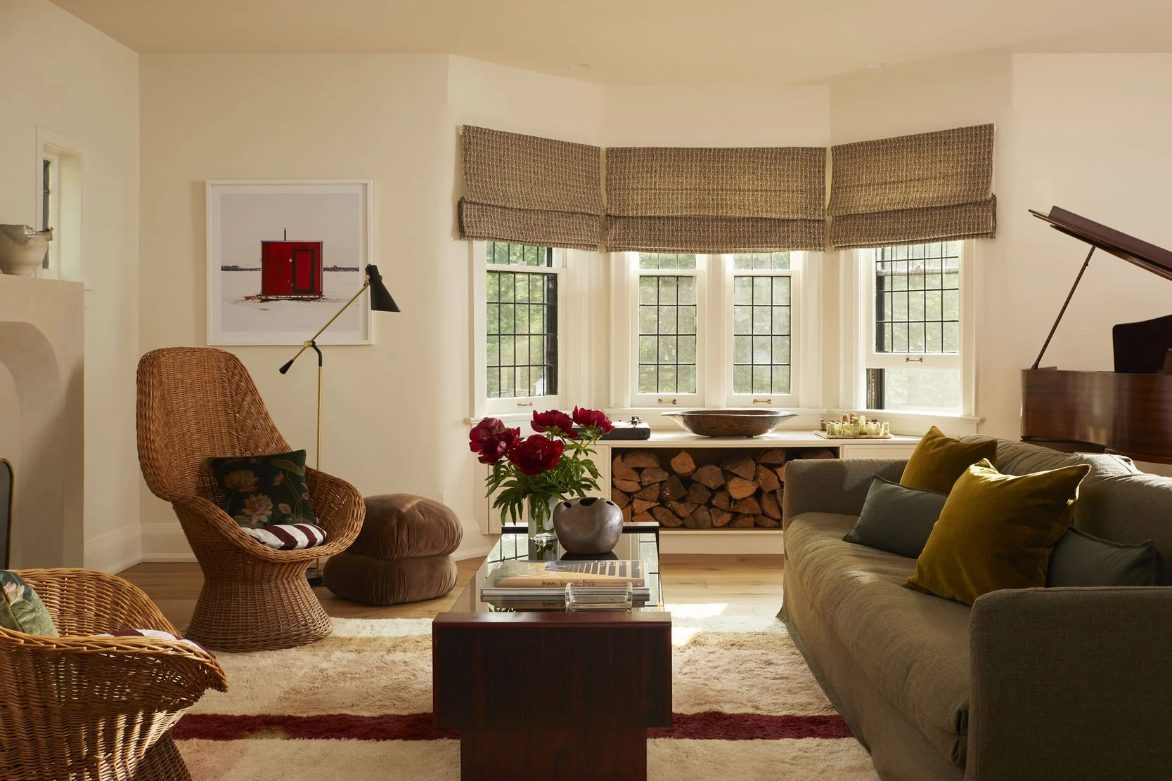 Living room with a green sofa, wicker armchairs, a coffee table with books and a vase of flowers, a fireplace with logs, and a bay window with a wood bowl on the sill.