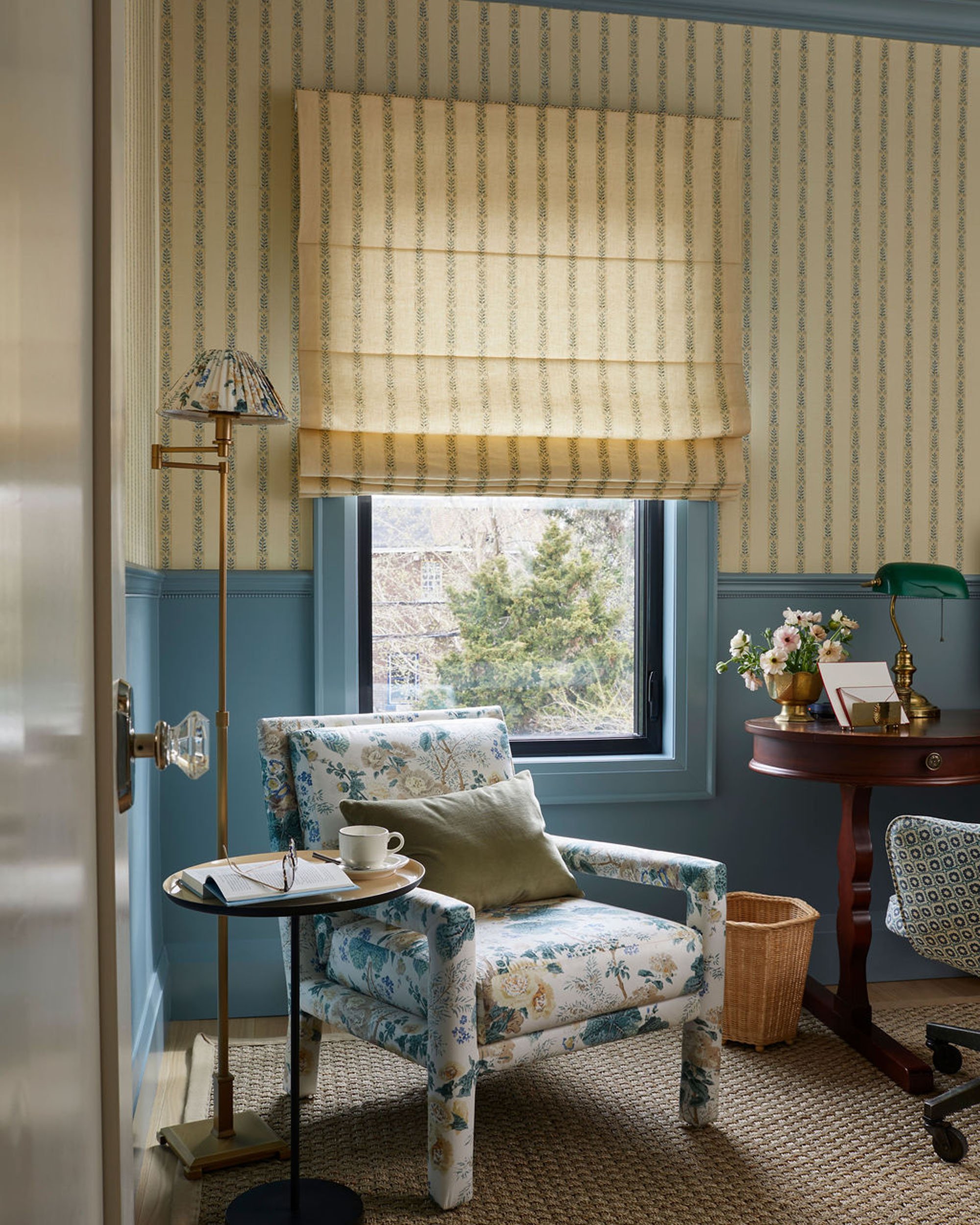 A cozy corner of a room featuring a floral armchair with a green pillow, a small round side table with a book, glasses, a cup, and a saucer, a floor lamp, a window with a beige Roman shade, a wooden desk with a green banker lamp, a flower vase, and a