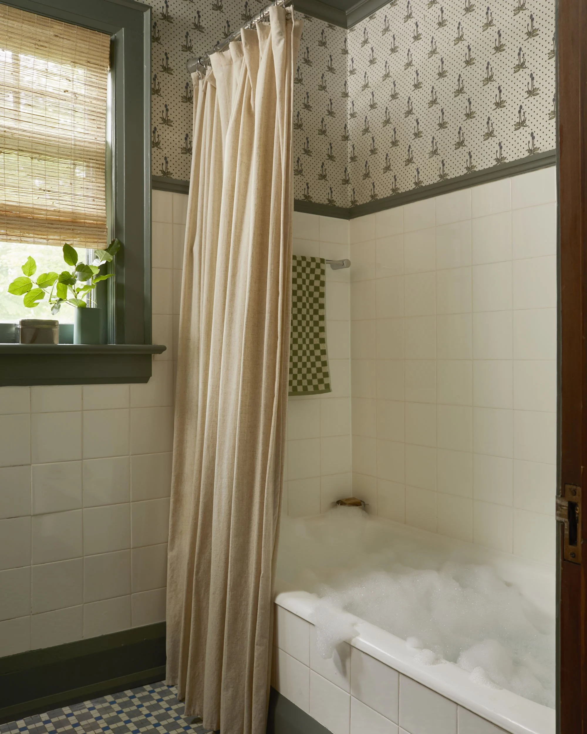 Bathroom with a bathtub filled with bubbles, beige shower curtain, window with bamboo blinds, patterned wallpaper with cacti, checkered towel hanging on the wall, wall tiles, and green trim.