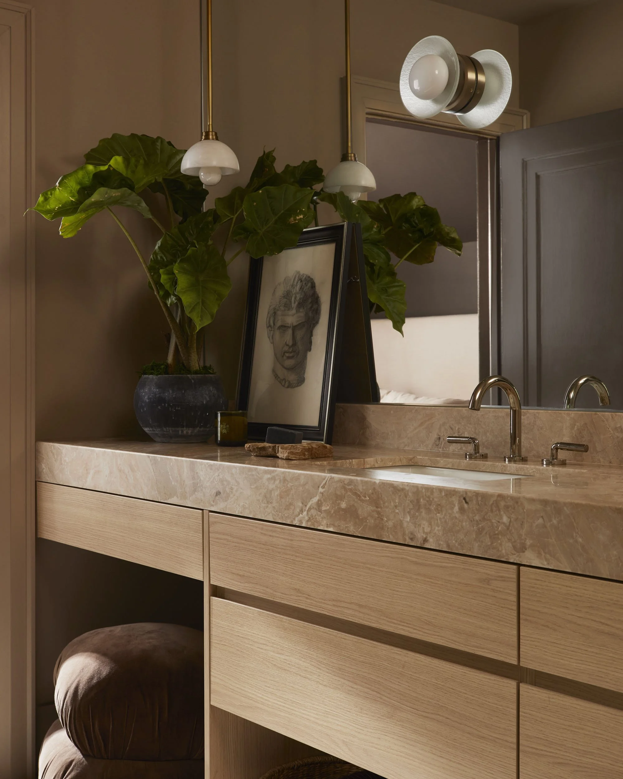 A bathroom vanity with a marble countertop, a framed sketch of a man's face, a large potted plant with big green leaves, and a small black object. Above, two hanging pendant lights and a modern wall sconce are visible. A large mirror reflects part of