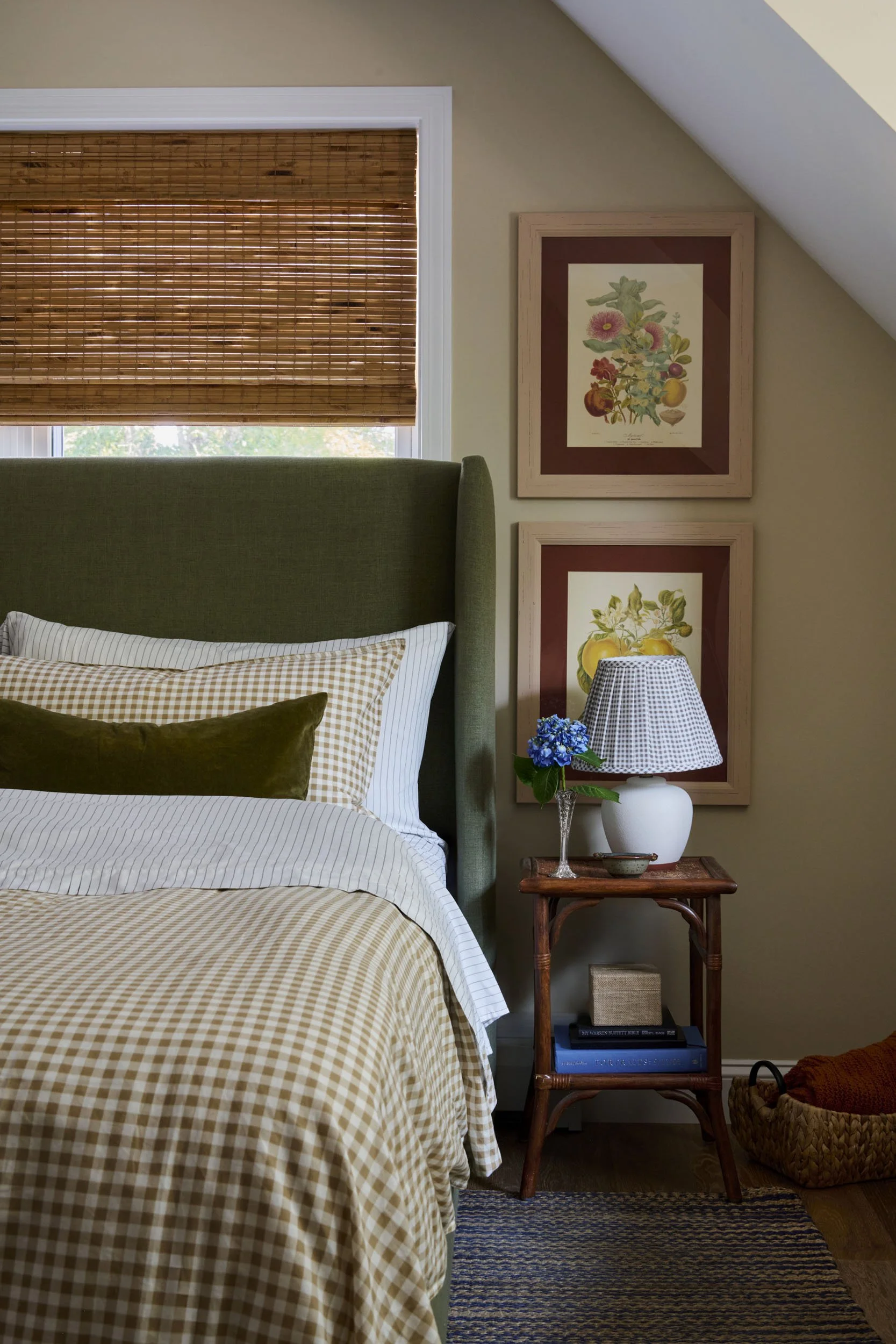 A cozy bedroom interior with a green upholstered bed, plaid bedding, two framed botanical prints on a beige wall, a wooden side table with a white vase, a checkered lampshade, a blue hydrangea, and a basket with orange blankets.