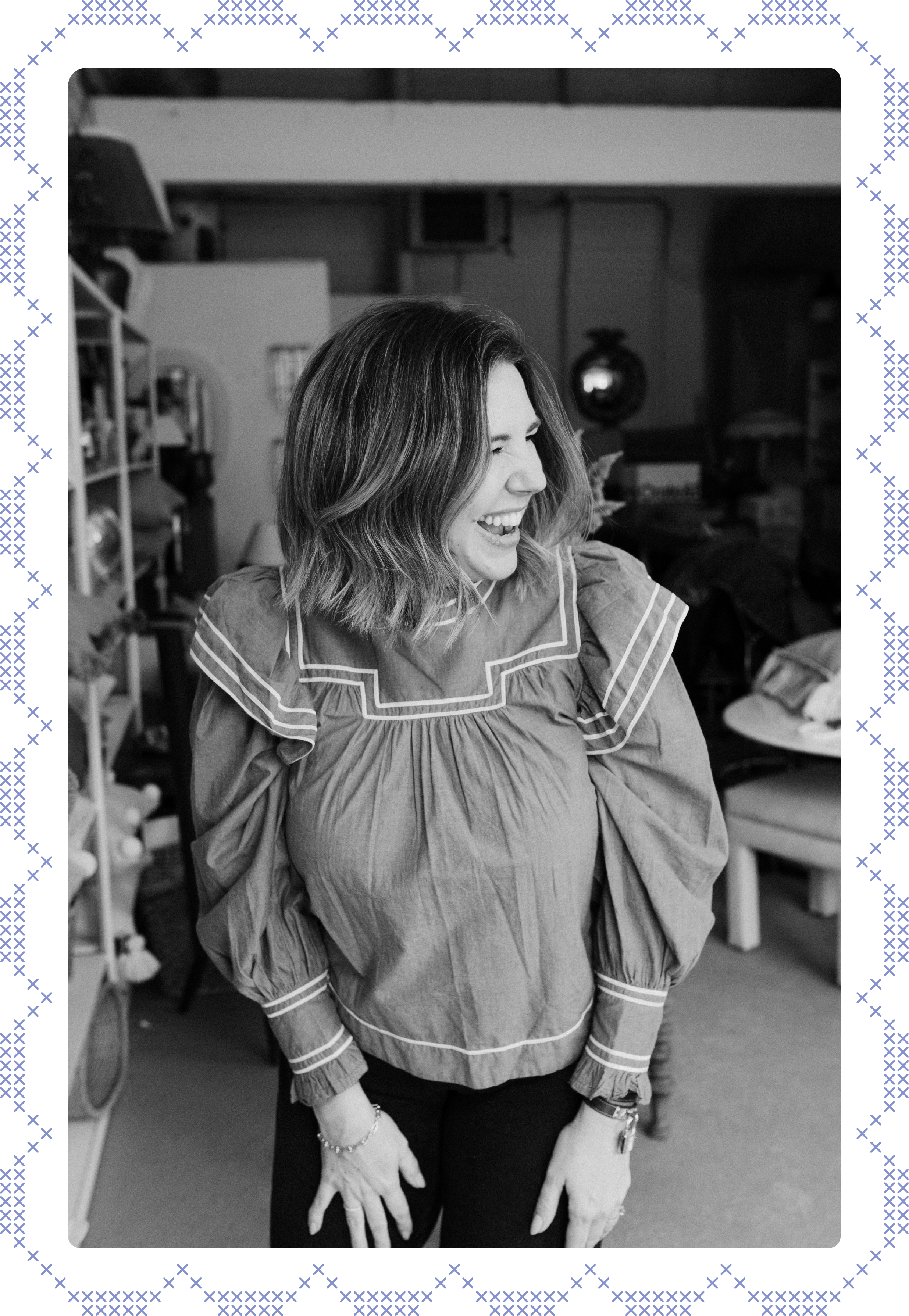 Kelly Lynn Armstrong smiling and looking to her right in an indoor setting, wearing a light-colored blouse with decorative stitching, standing with hands on her thighs.