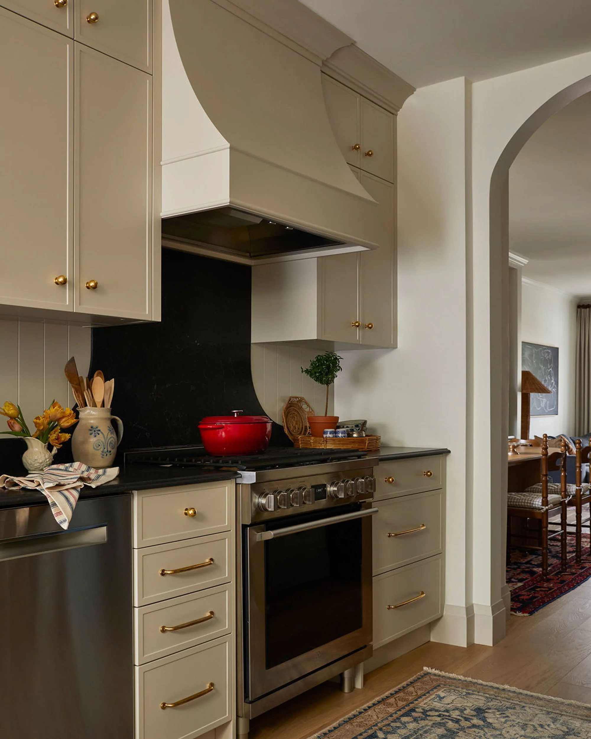 Kitchen with cream-colored cabinets with gold knobs, black countertop, stainless steel oven, red pot on stove, decorative items including a small potted plant and flowers, doorway leading to a dining area with a rug and wooden chairs.