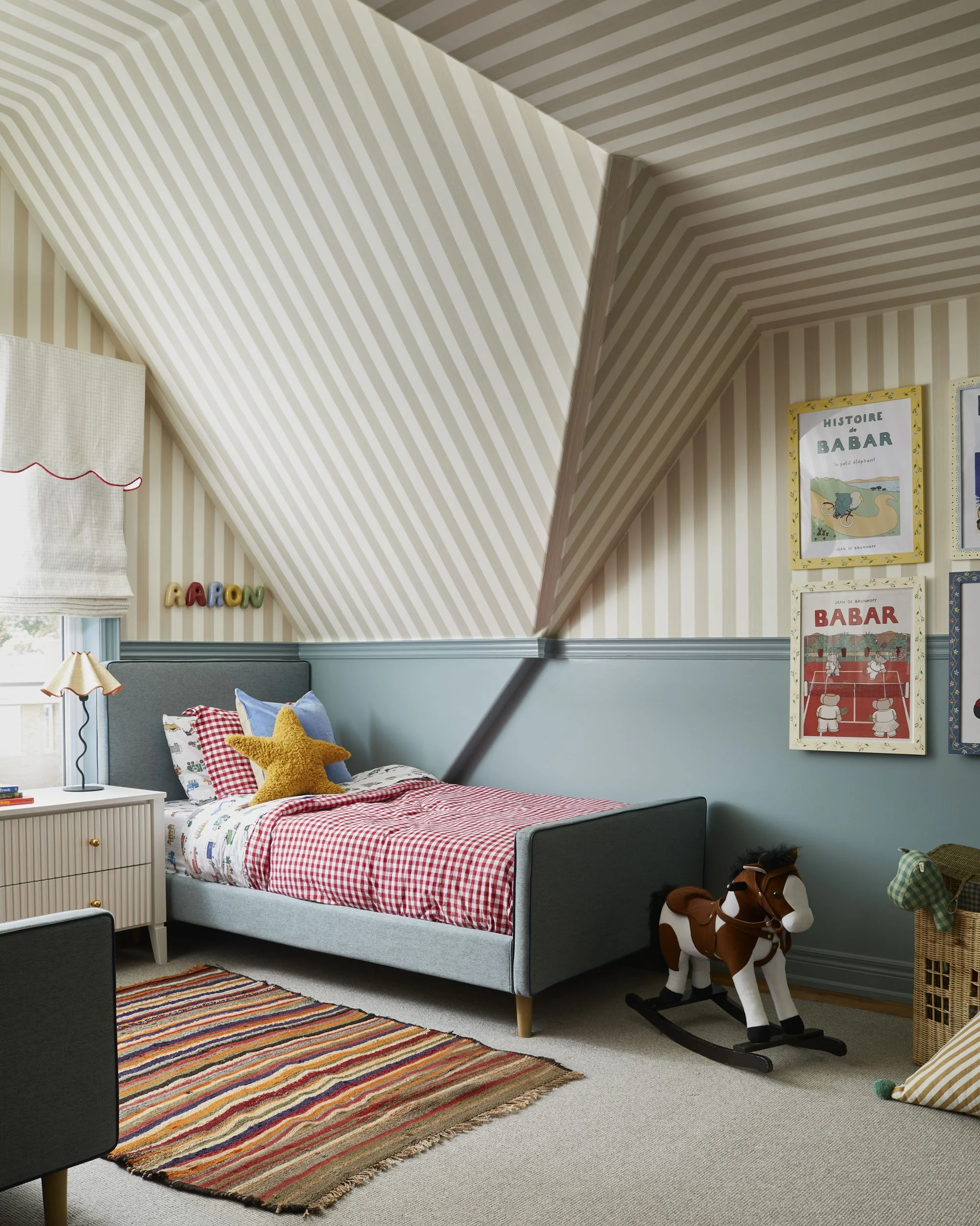 Decorated children's bedroom featuring a bed with red and white checkered bedding, colorful pillows, a star-shaped pillow, a striped area rug, a rocking horse, framed children's book posters on the wall, a bedside table, and a window with a white shade.