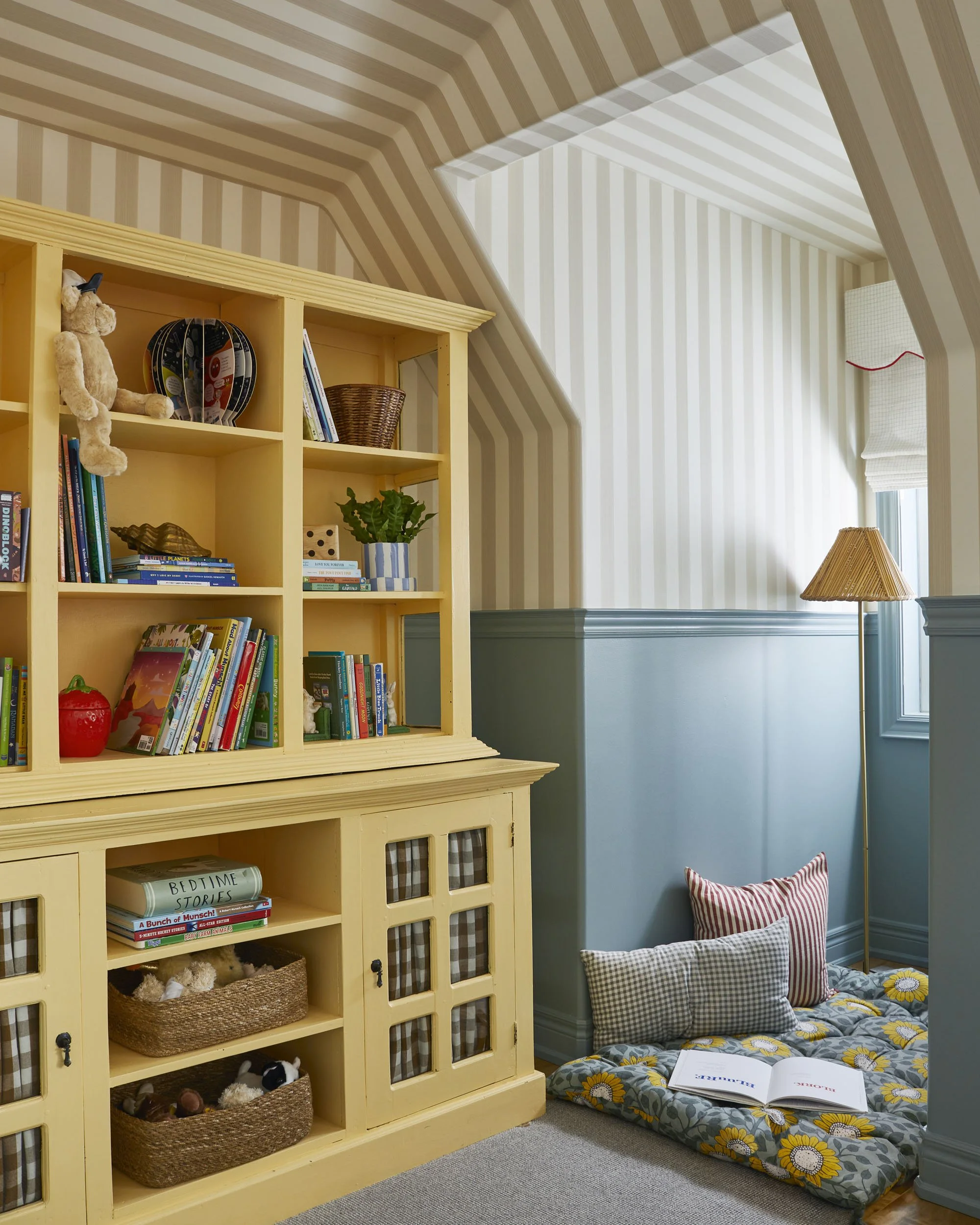 A cozy corner of a room with a yellow bookshelf filled with books, toys, and decorative items, next to a reading nook with pillows and an open book, under a window with a white shade.