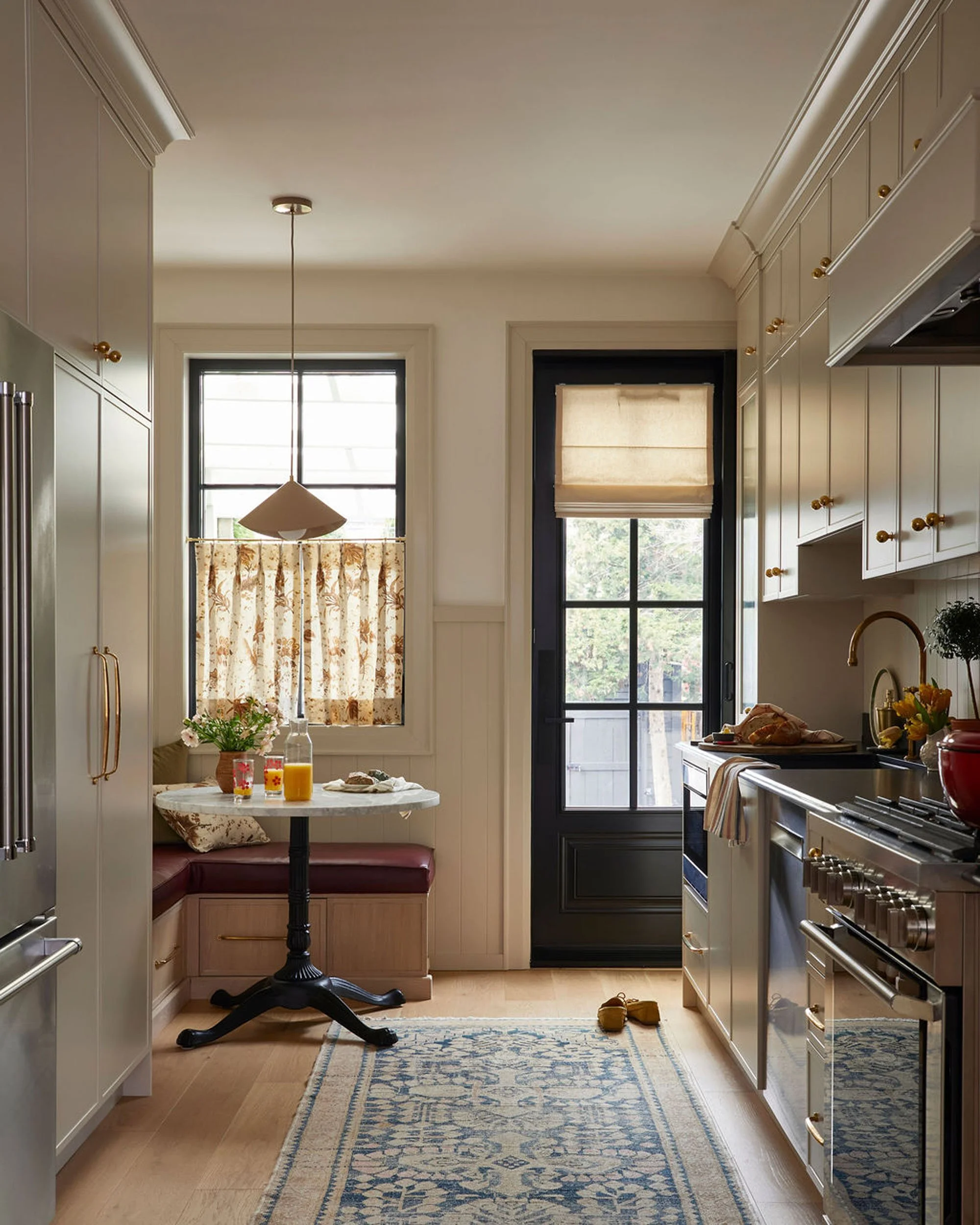 Sunlit kitchen with white cabinets, a small breakfast nook with a round table and built-in bench, a stove, a sink, and a black door leading outside. There are breakfast items and a potted plant on the counter.