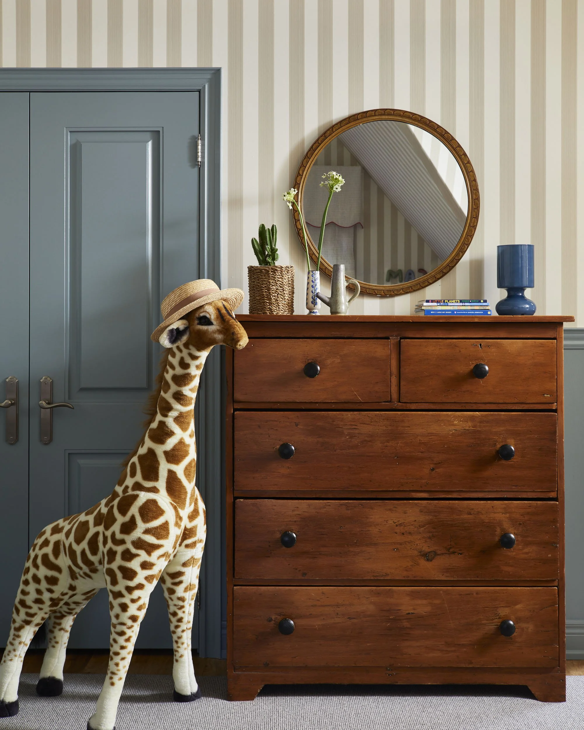 A room featuring a wooden dresser with a mirror above it. Decor includes a cactus plant, a tall white flower in a vase, a small watering can, and a blue lamp. A stuffed giraffe wearing a hat stands beside the dresser.