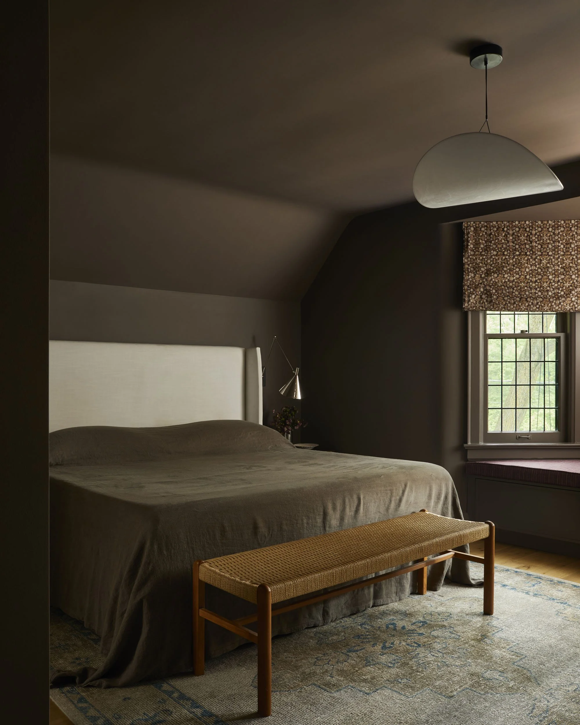 A bedroom with a large bed, beige bedding, and a woven bench at the foot. There is a window with a floral-patterned curtain, a built-in window seat, and modern wall lamps. The room has dark-colored walls and a ceiling with a hanging light fixture.
