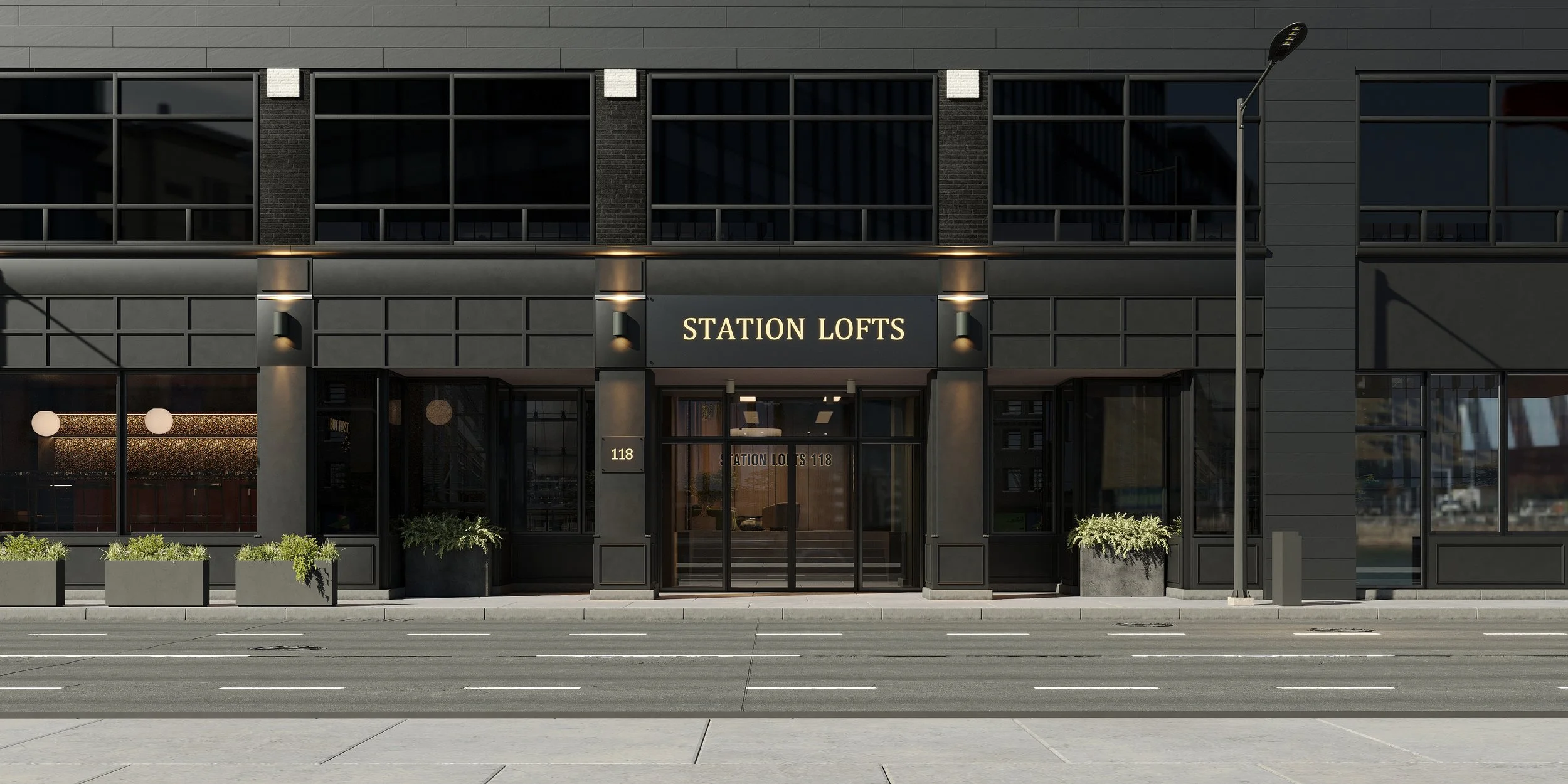 Modern building with a sign that reads 'Station Lofts' on the facade, large glass windows, black exterior, outdoor sidewalk with planters, streetlights, and parking lines visible in the foreground.